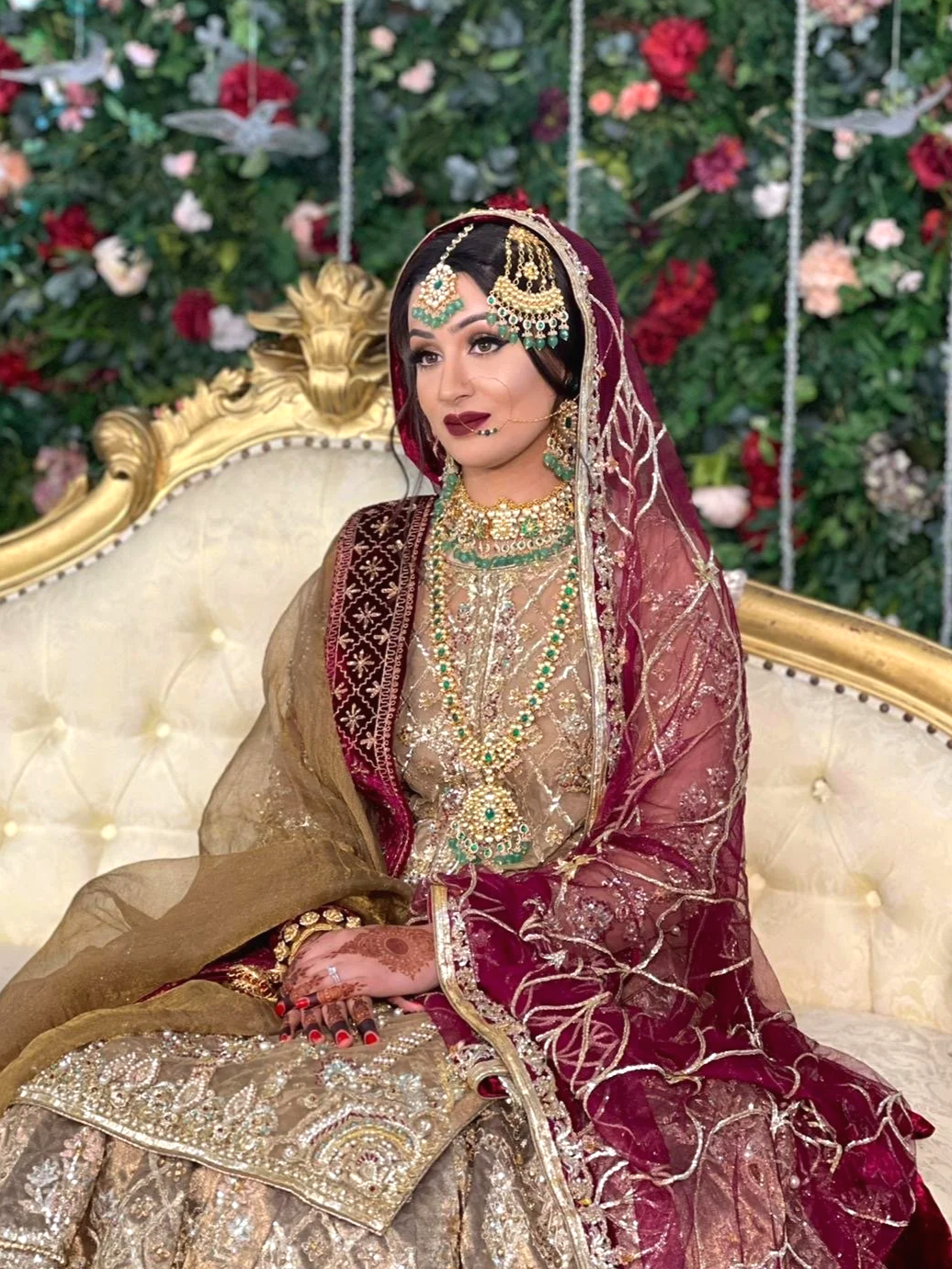 A woman dressed in traditional Indian bridal attire, wearing elaborate jewelry and a maroon and gold embroidered sari, sitting on a luxurious cream-colored sofa with a floral backdrop.