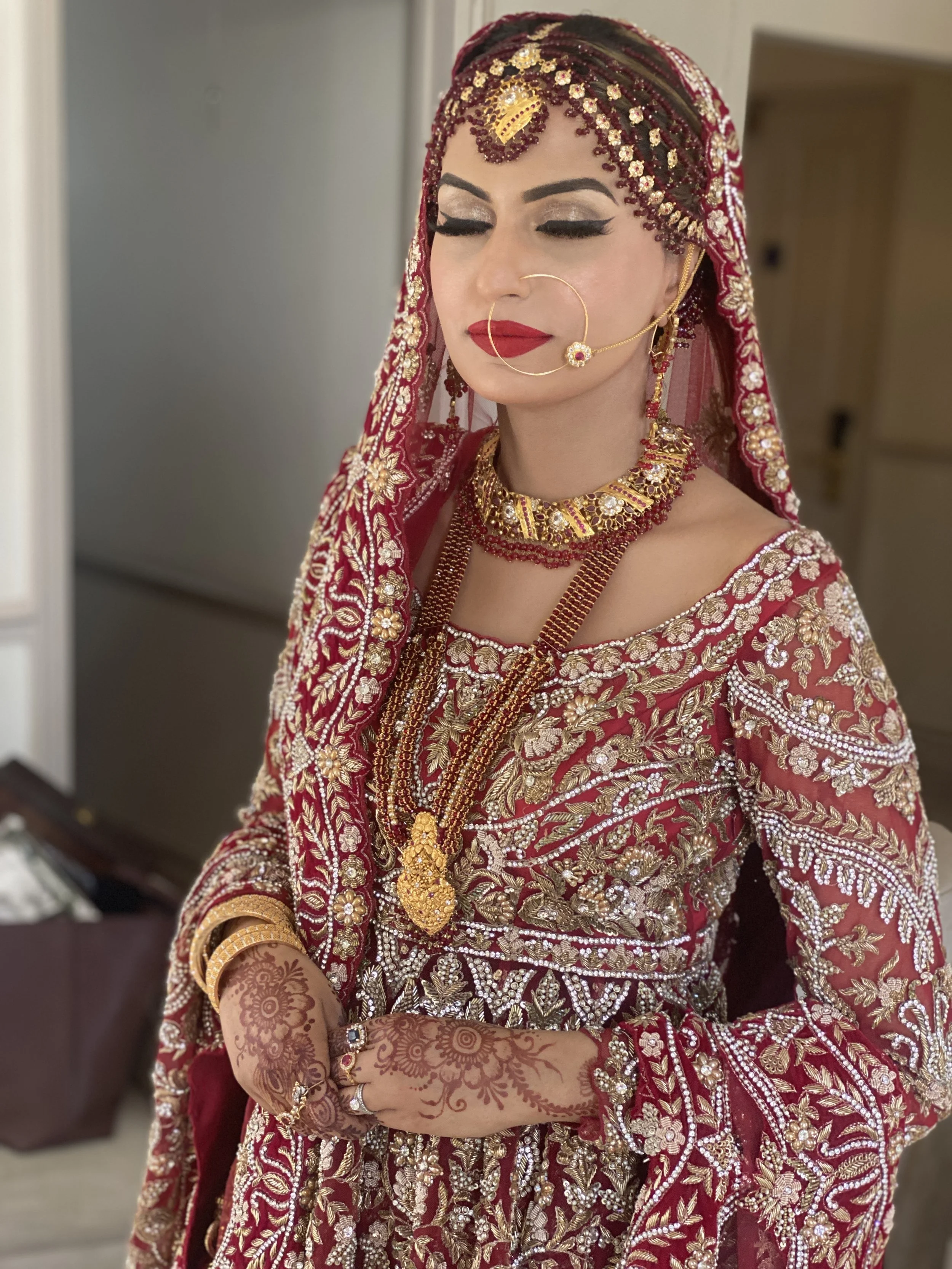 A woman dressed in traditional Indian wedding attire, wearing a heavily embroidered red and gold saree with matching jewelry, henna on her hands, and traditional accessories including a nose ring and necklace, standing with eyes closed.