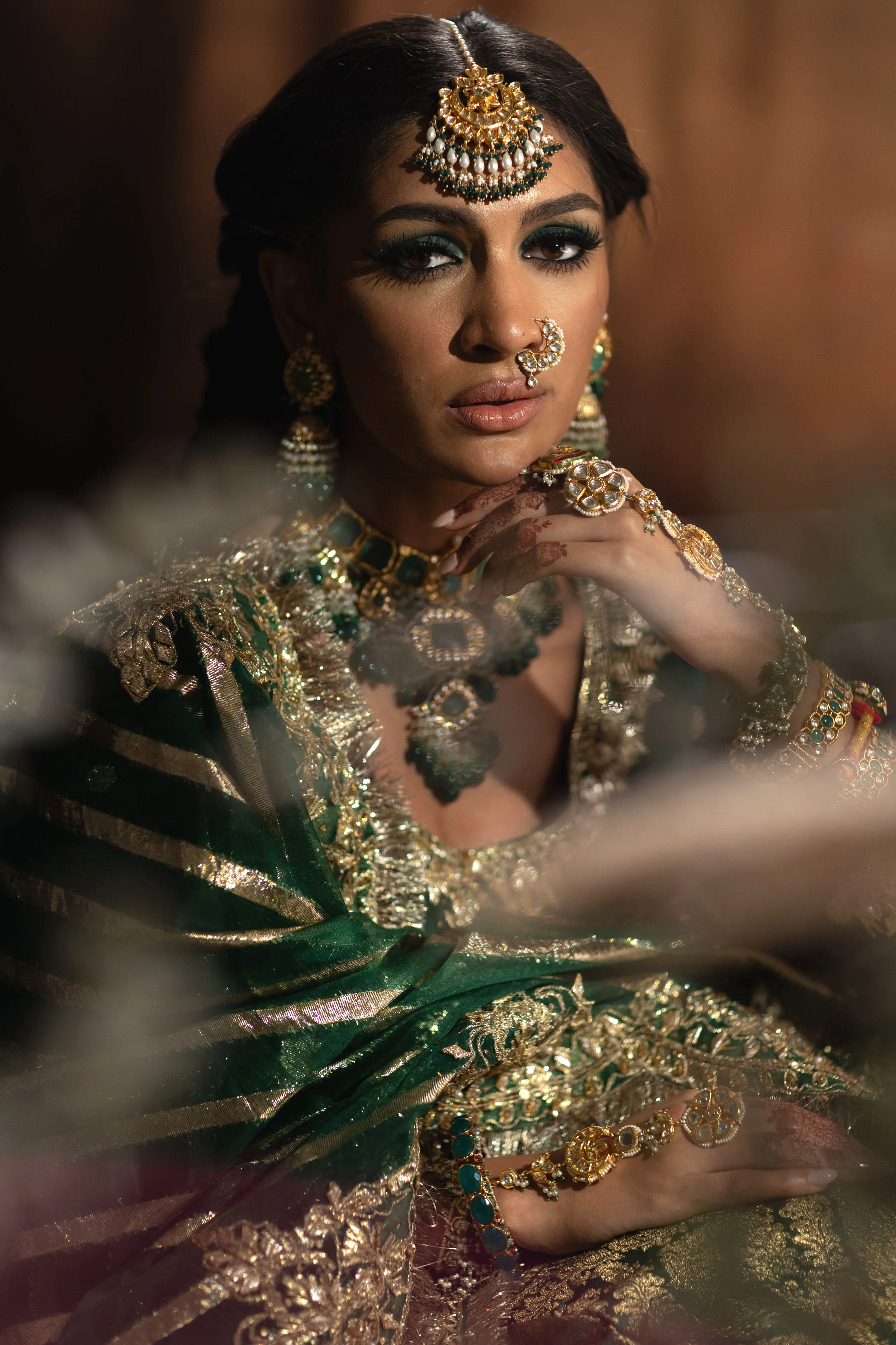 A woman dressed in traditional Indian attire and heavy jewelry, including a nose ring, earrings, necklace, and headpiece, sitting elegantly with her hand resting near her chin.