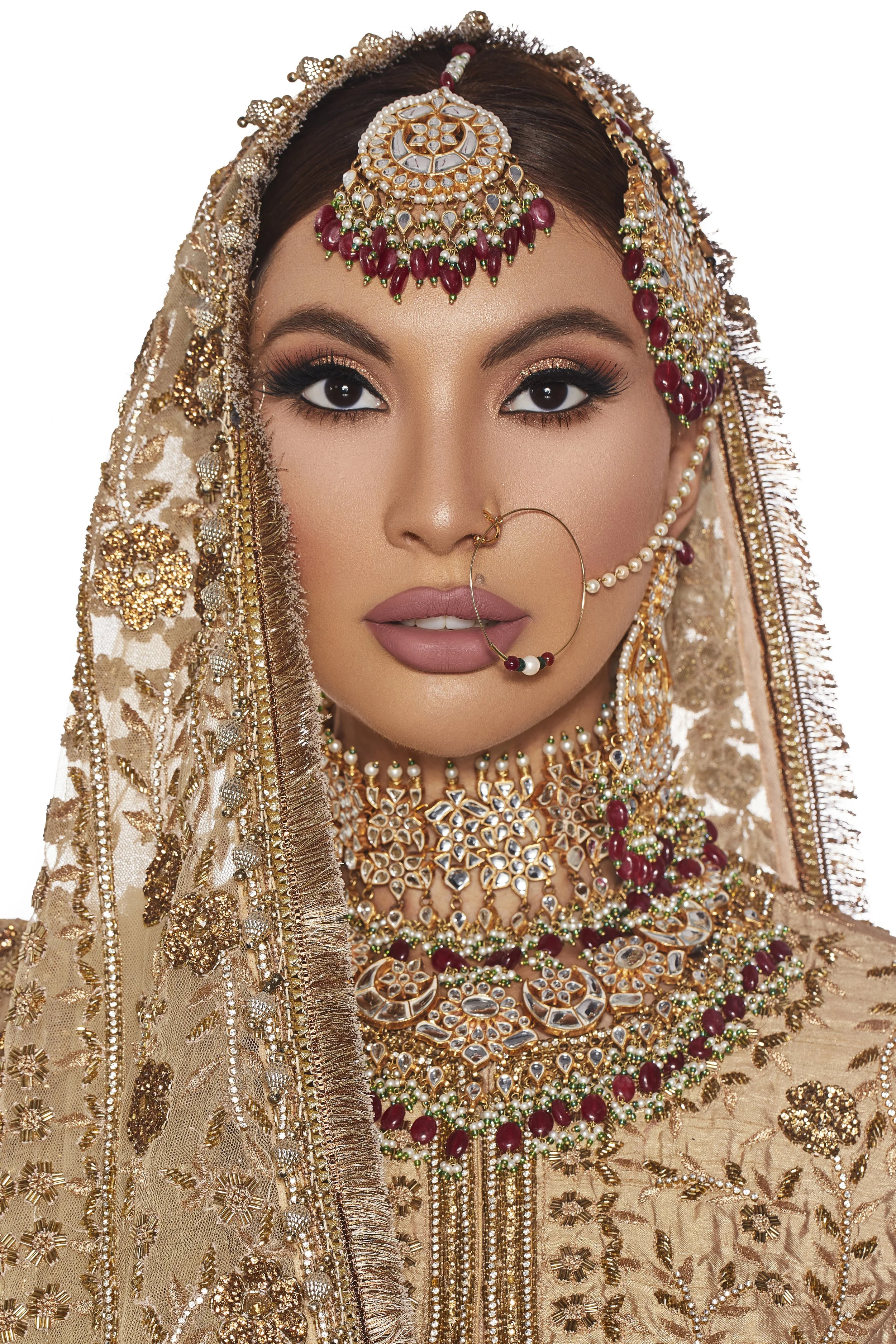 A woman dressed in traditional Indian bridal attire adorned with elaborate gold jewelry, including a headpiece, necklace, earrings, nose ring, and heavily embroidered fabric.