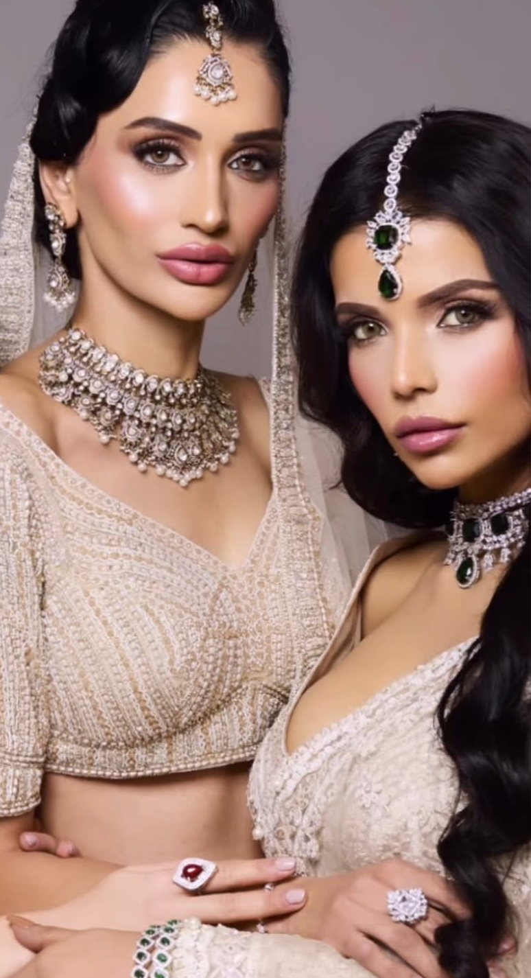 Two women dressed in traditional Indian attire with elaborate jewelry, including necklaces, earrings, and headpieces. One woman has dark, wavy hair, and both have makeup with accentuated eyes and lips.