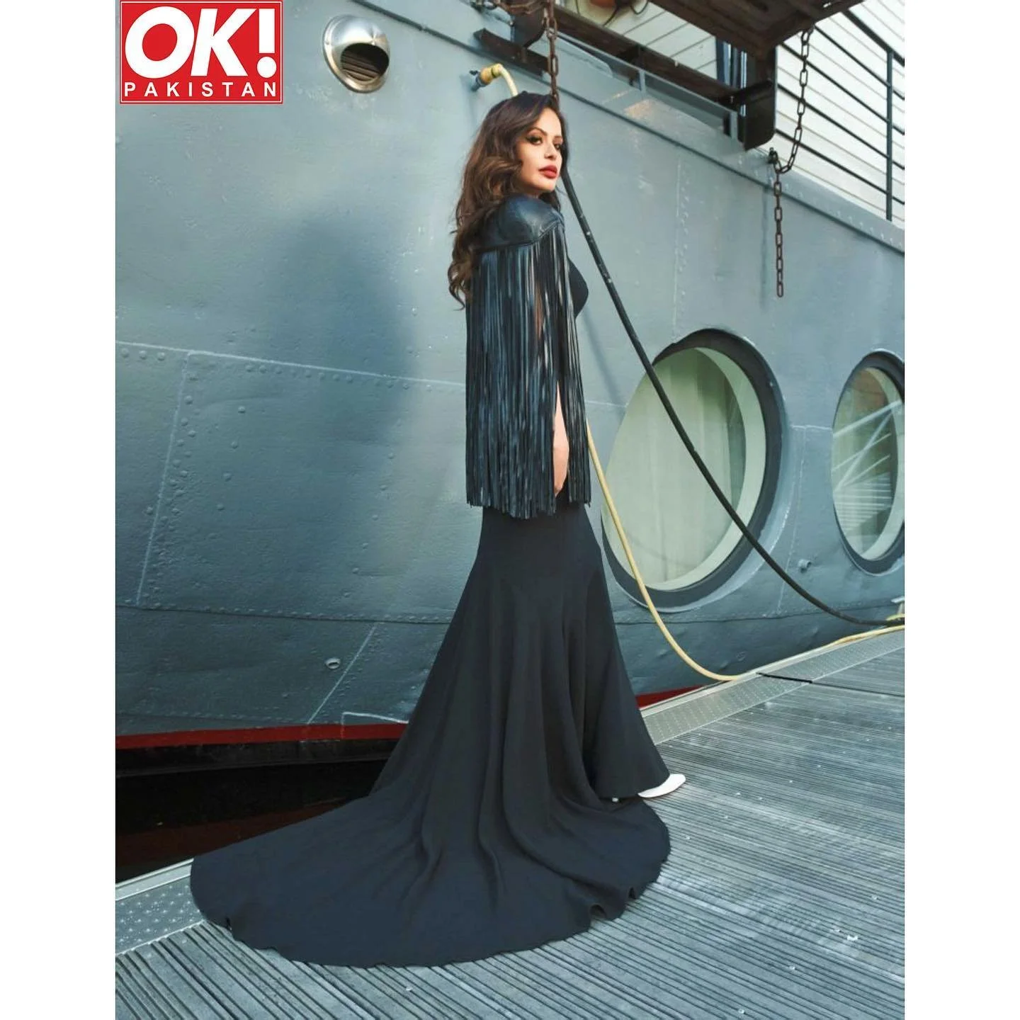 A woman in a long black gown and black leather jacket with fringe detail stands on a ship's deck, looking at the camera with a serious expression, against a gray ship wall with round windows and industrial equipment.