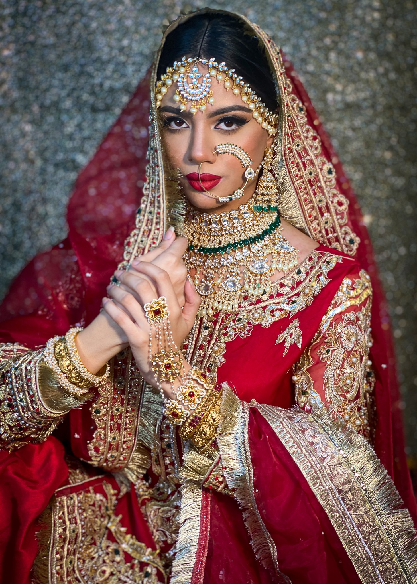 A woman dressed in traditional Indian bridal attire, wearing a red and gold embroidered saree, adorned with multiple layers of gold and gemstone jewelry, including necklaces, earrings, and a headpiece.