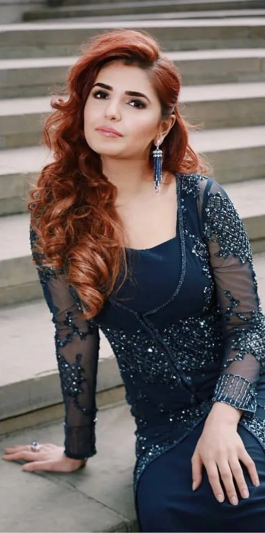 A woman with long, wavy red hair sitting on stairs in a dark blue sequined dress with sheer sleeves and earrings.