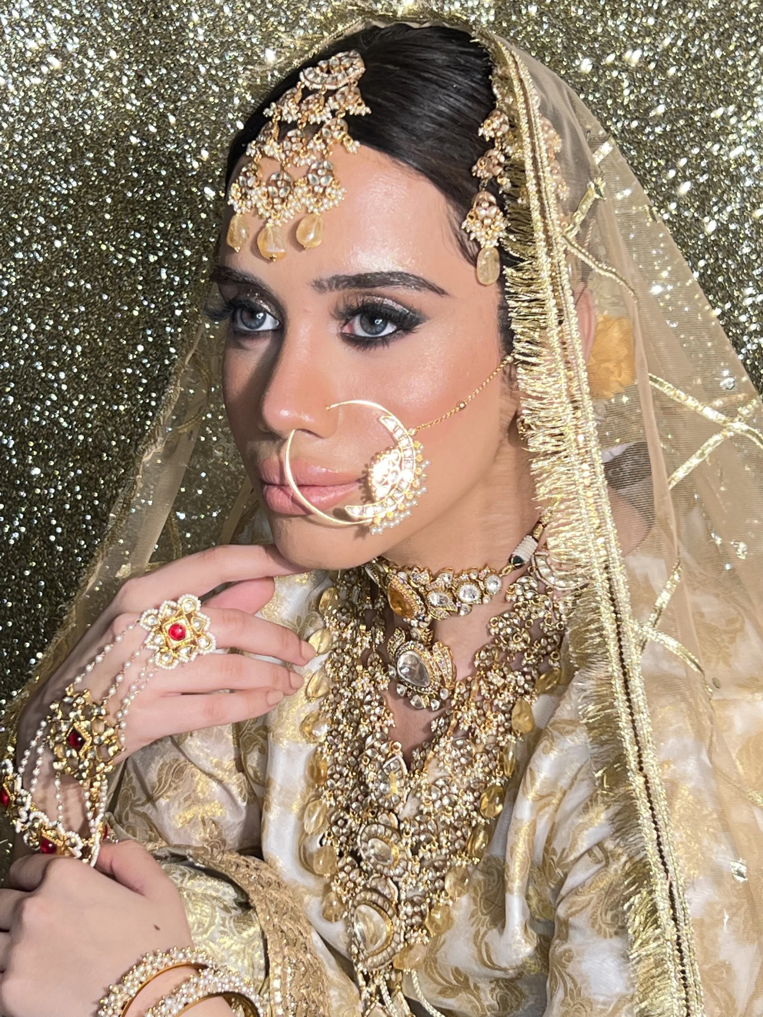 A woman dressed in traditional Indian bridal attire, adorned with gold jewelry, including a headpiece, nose ring, necklaces, rings, and bangles, with a gold embroidered veil and a gold glittery background.