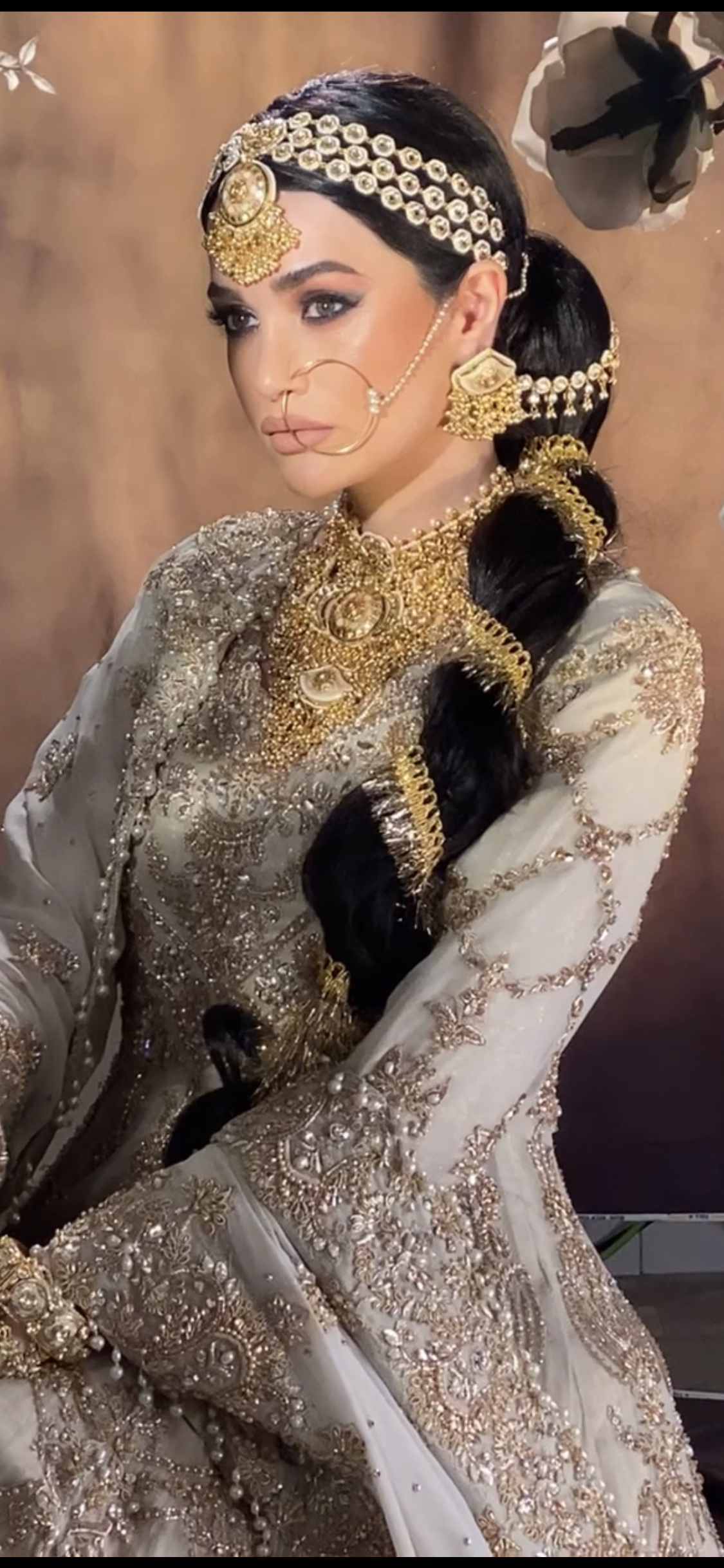 A woman dressed in traditional Indian bridal attire, wearing ornate gold jewelry including a headpiece, nose ring, earrings, necklace, and arm jewelry, with a long black braid decorated with gold accessories.