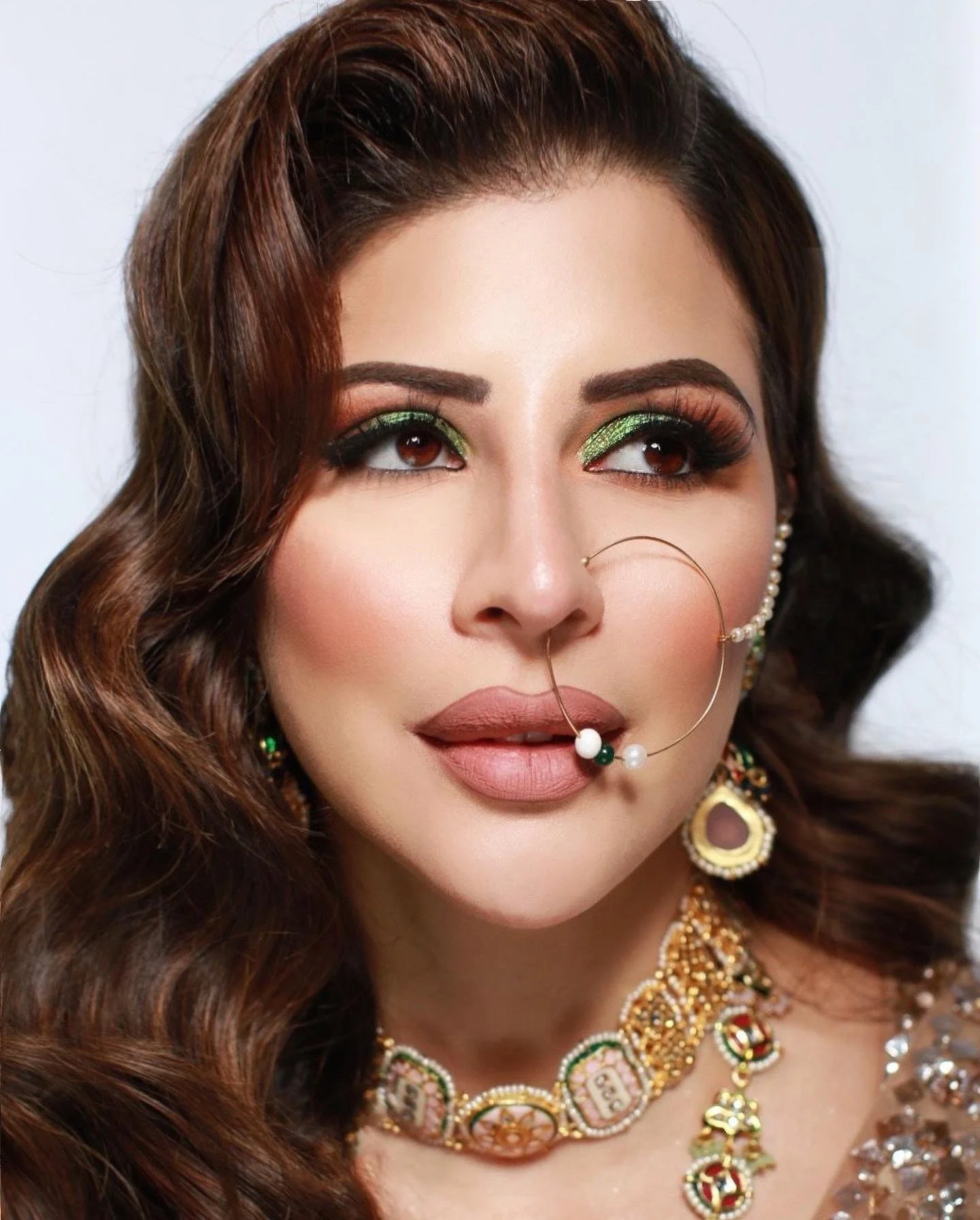 Close-up of a woman with wavy brown hair, wearing bright green eye shadow, pearl and gold jewelry, and a decorative nose ring.
