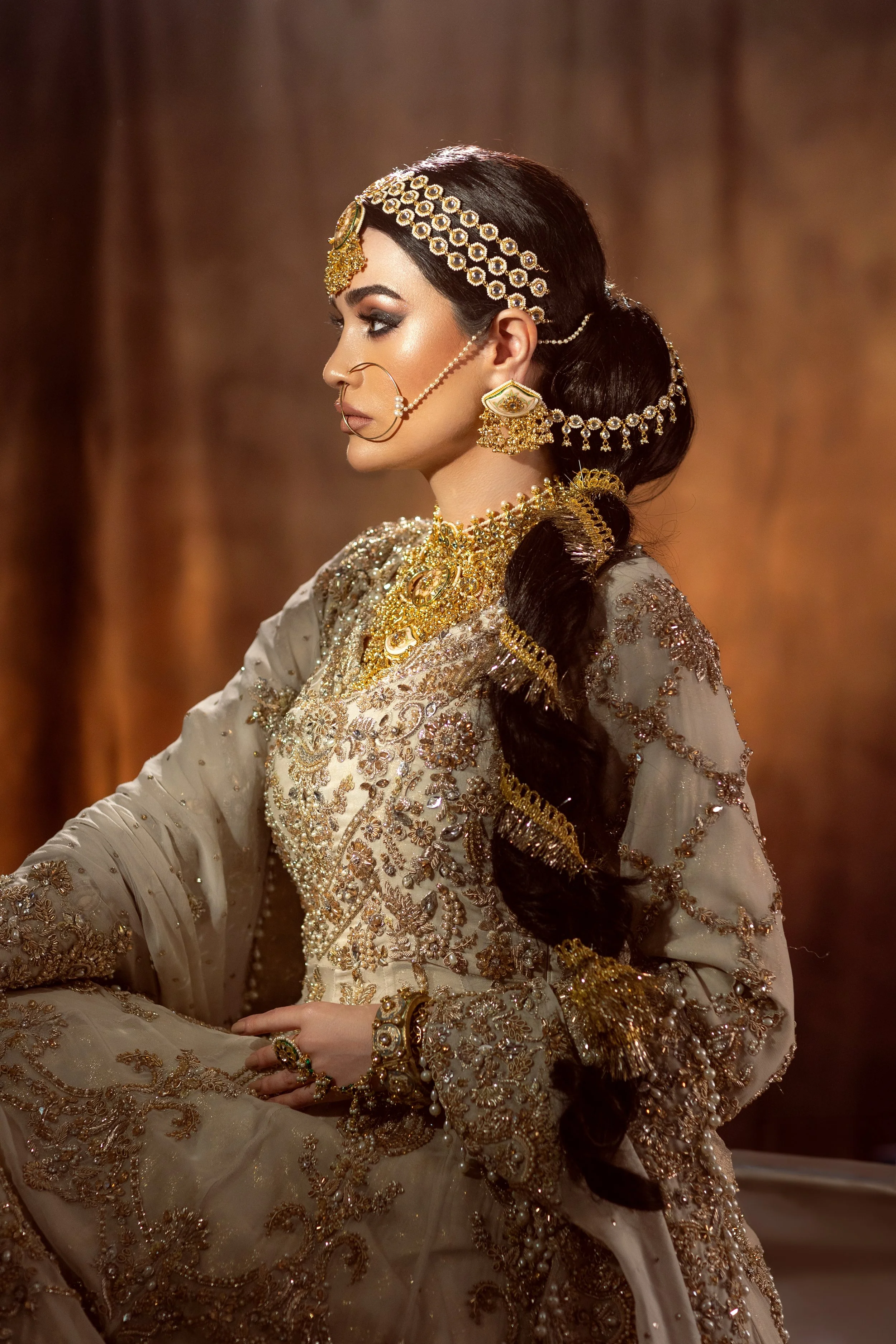 A woman dressed in traditional Indian bridal attire, heavily adorned with gold jewelry and embroidery, sitting in profile against a warm background.