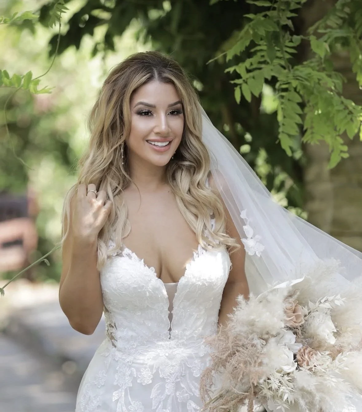 A smiling bride in a white lace wedding gown with a deep V neckline, holding a bouquet of neutral-colored flowers, standing outdoors with green foliage in the background.