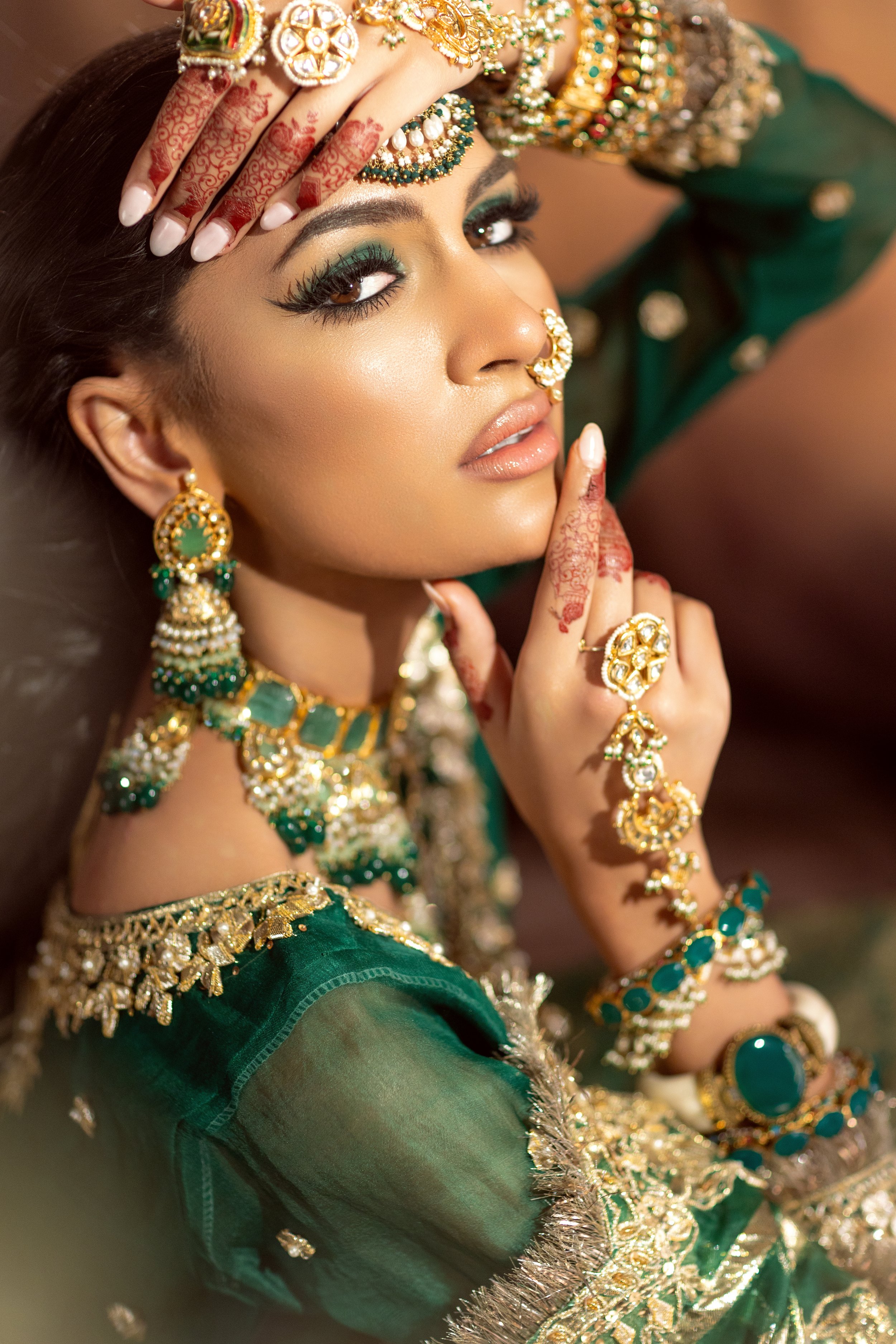 A woman dressed in traditional Indian attire, adorned with gold jewelry including earrings, necklaces, bangles, and headpieces, and decorated with intricate henna designs on her hands and fingers, posing with her hand on her forehead and chin.