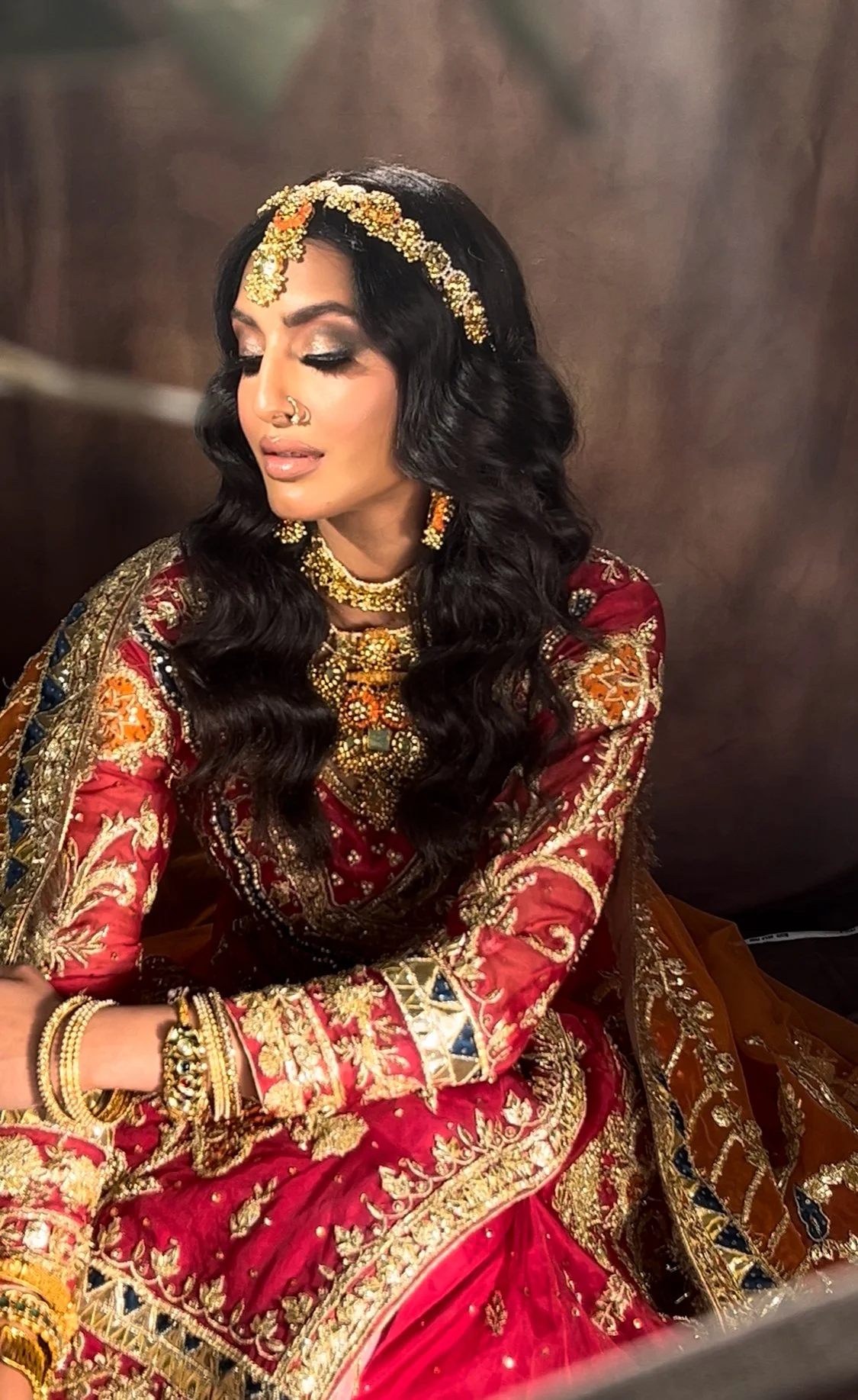 A woman dressed in traditional South Asian bridal attire, with intricate gold jewelry, a red embroidered dress, and a decorative headpiece, sitting with her eyes closed.