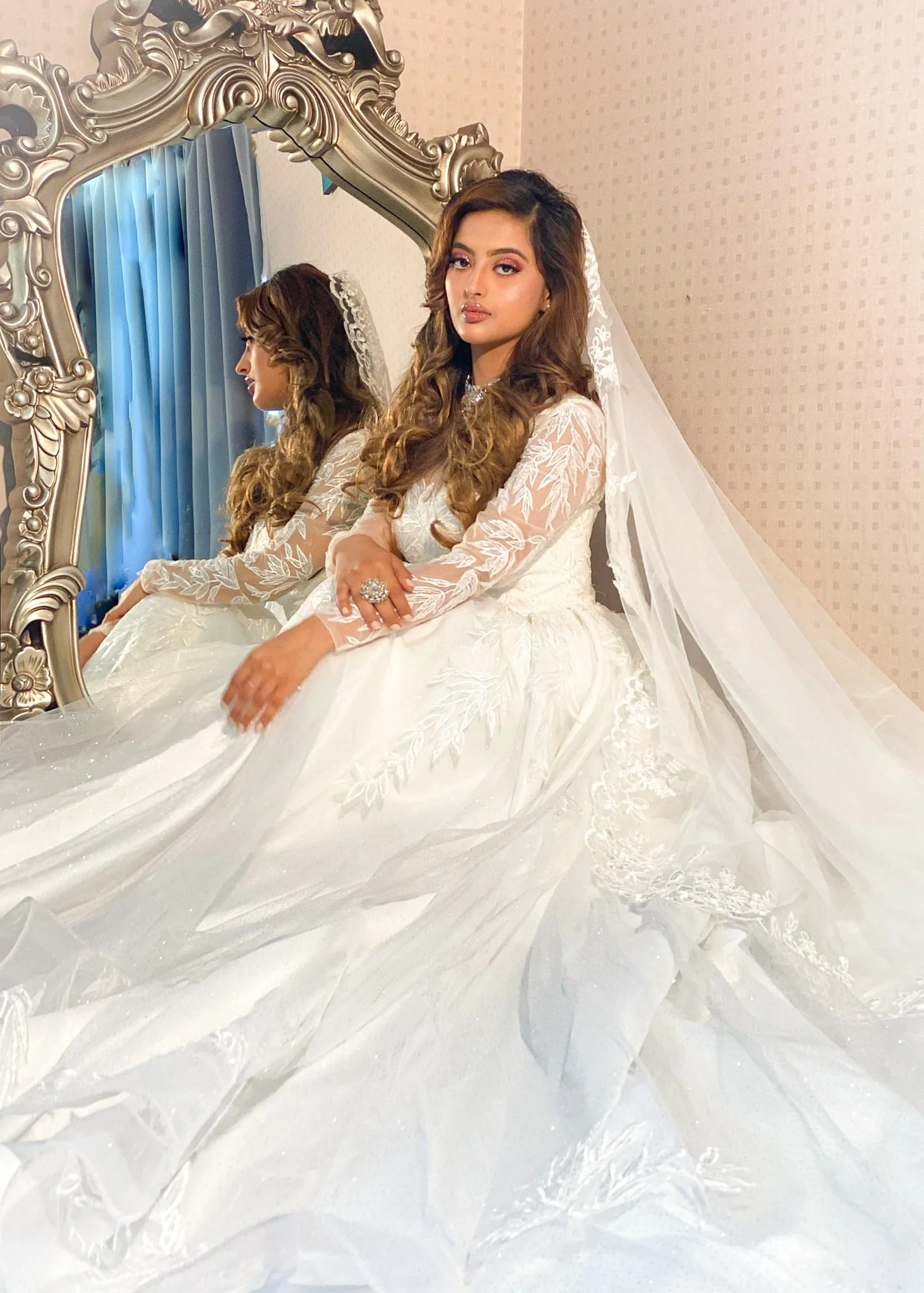 A woman in a white wedding dress with lace and floral embroidery sits in front of an ornate mirror, wearing a veil and a large ring, with a reflection showing her profile. The room has cream-colored walls and blue curtains.
