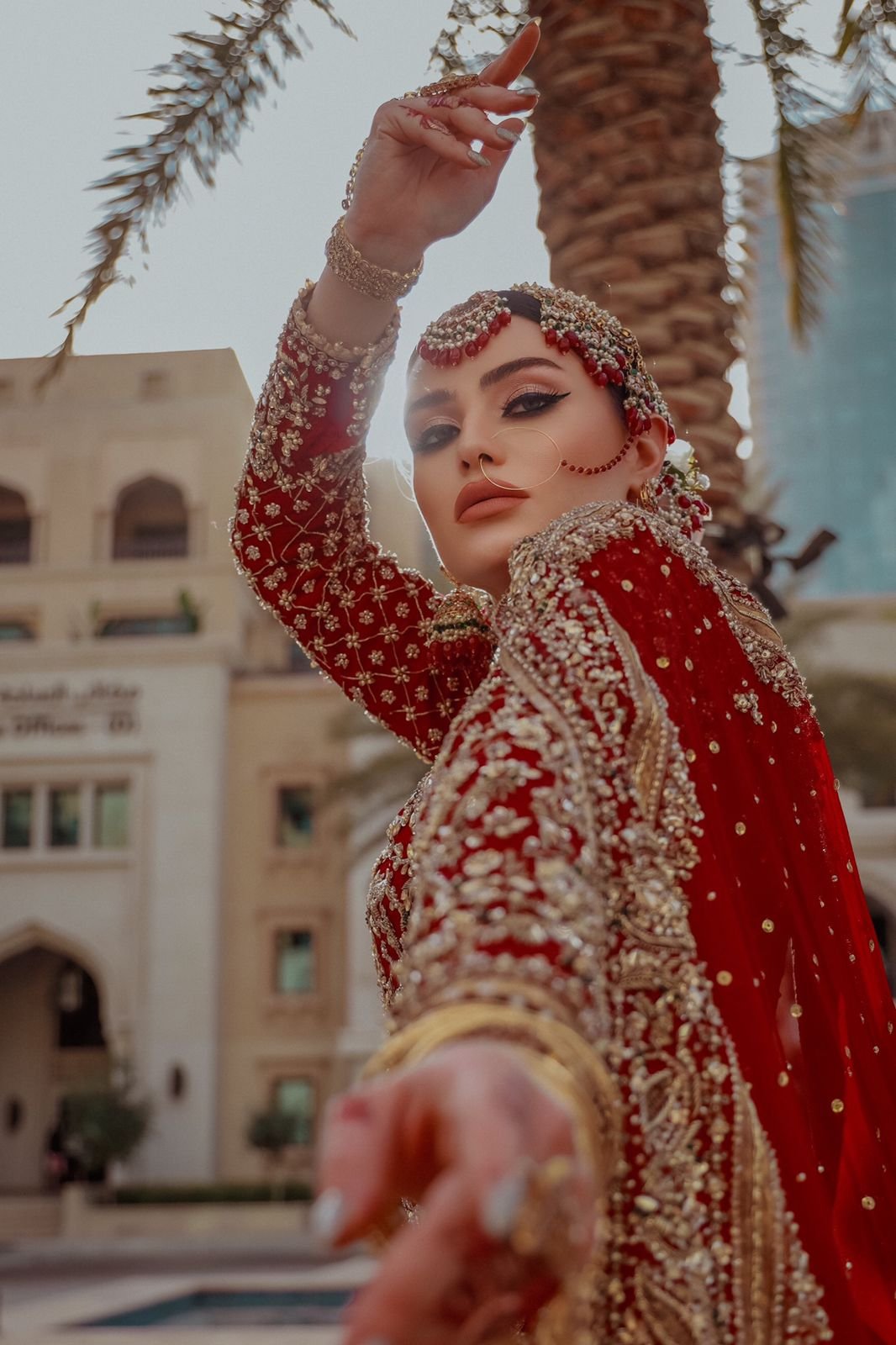 A woman dressed in traditional South Asian bridal attire with intricate gold embroidery, jewelry, and a nose ring, posed outdoors next to a palm tree with a building in the background.