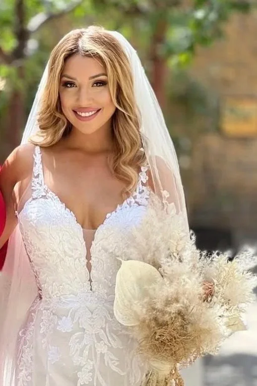 A smiling bride with blonde hair wearing a white wedding dress with lace details and a veil, holding a bouquet of dried pampas grass outdoors.