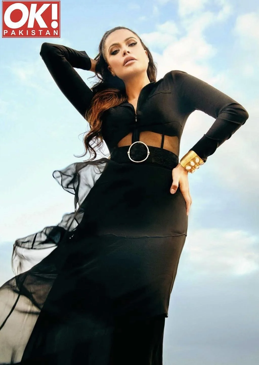 A woman in a black outfit with a sheer top and a long skirt poses outdoors against a blue sky with clouds, with her hand on her hip and one arm raised behind her head.