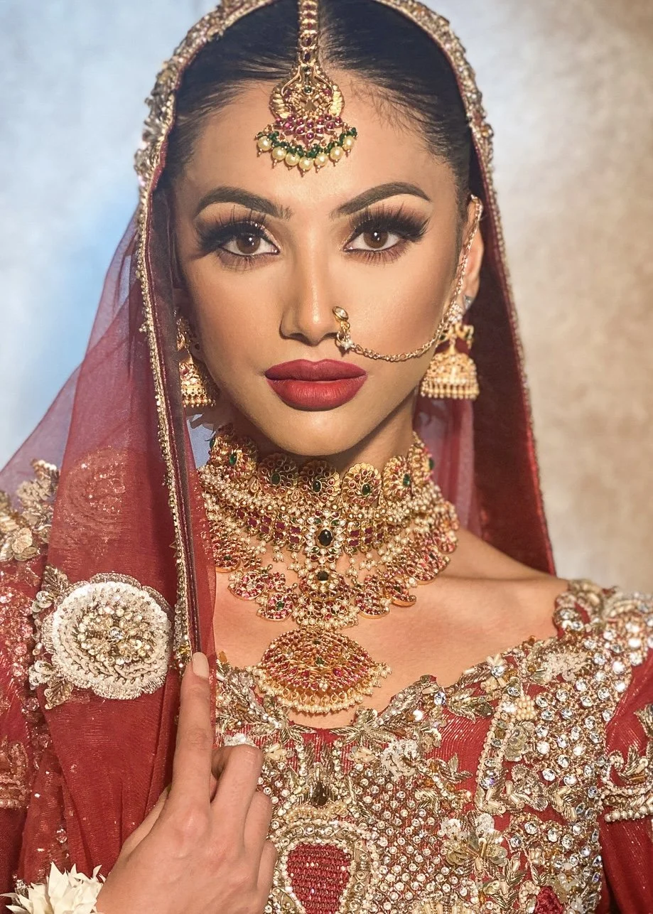 Close-up of a woman dressed in traditional Indian bridal attire, wearing elaborate gold and gemstone jewelry, a red saree with intricate embroidery, and a sheer red veil with gold embellishments.