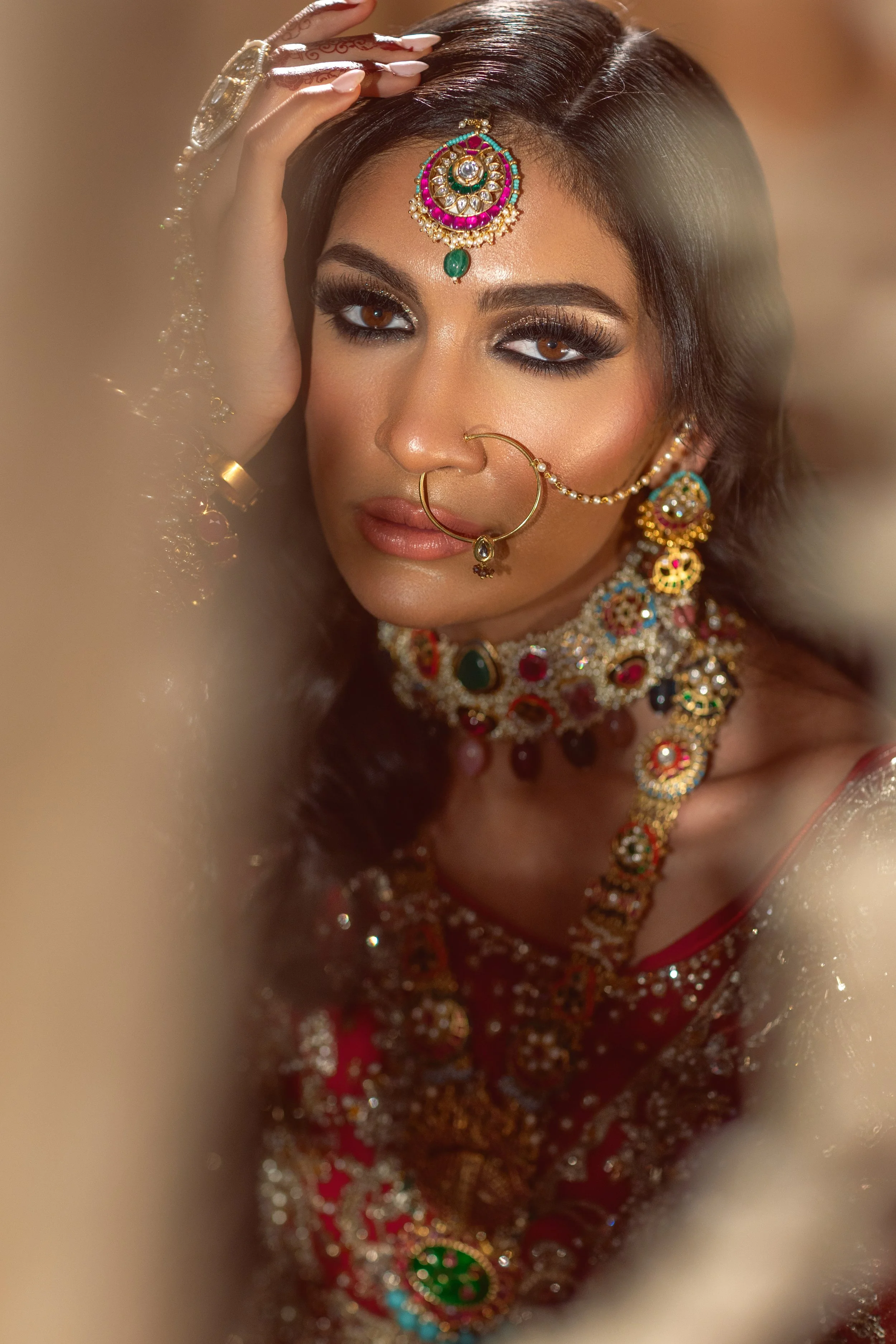 Close-up of a woman dressed in traditional Indian jewelry and attire, with ornate earrings, necklaces, and a nose ring. She has dark hair, makeup, and is looking at the camera with a soft expression.
