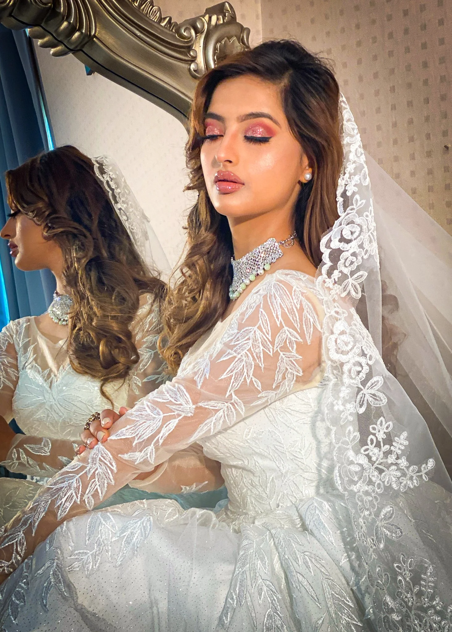 A woman dressed as a bride wearing a lace wedding dress with floral embroidery, veil, pearl earrings, and layered necklace, standing in front of a mirror with closed eyes, showing makeup and styled hair.