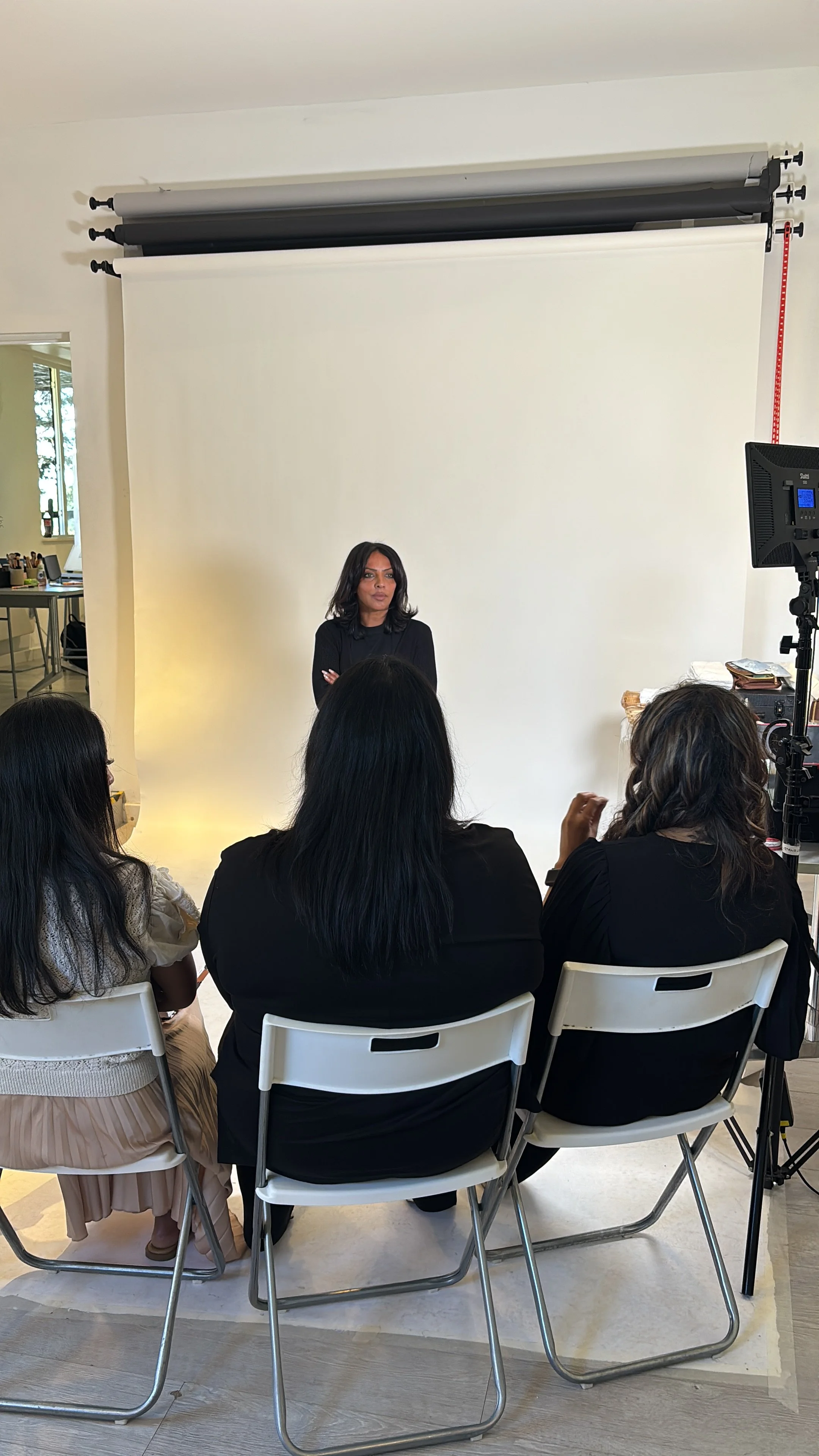 A woman in black clothes standing in front of a white backdrop during a presentation or event, with four women seated facing her, and a lighting and camera setup to the right.