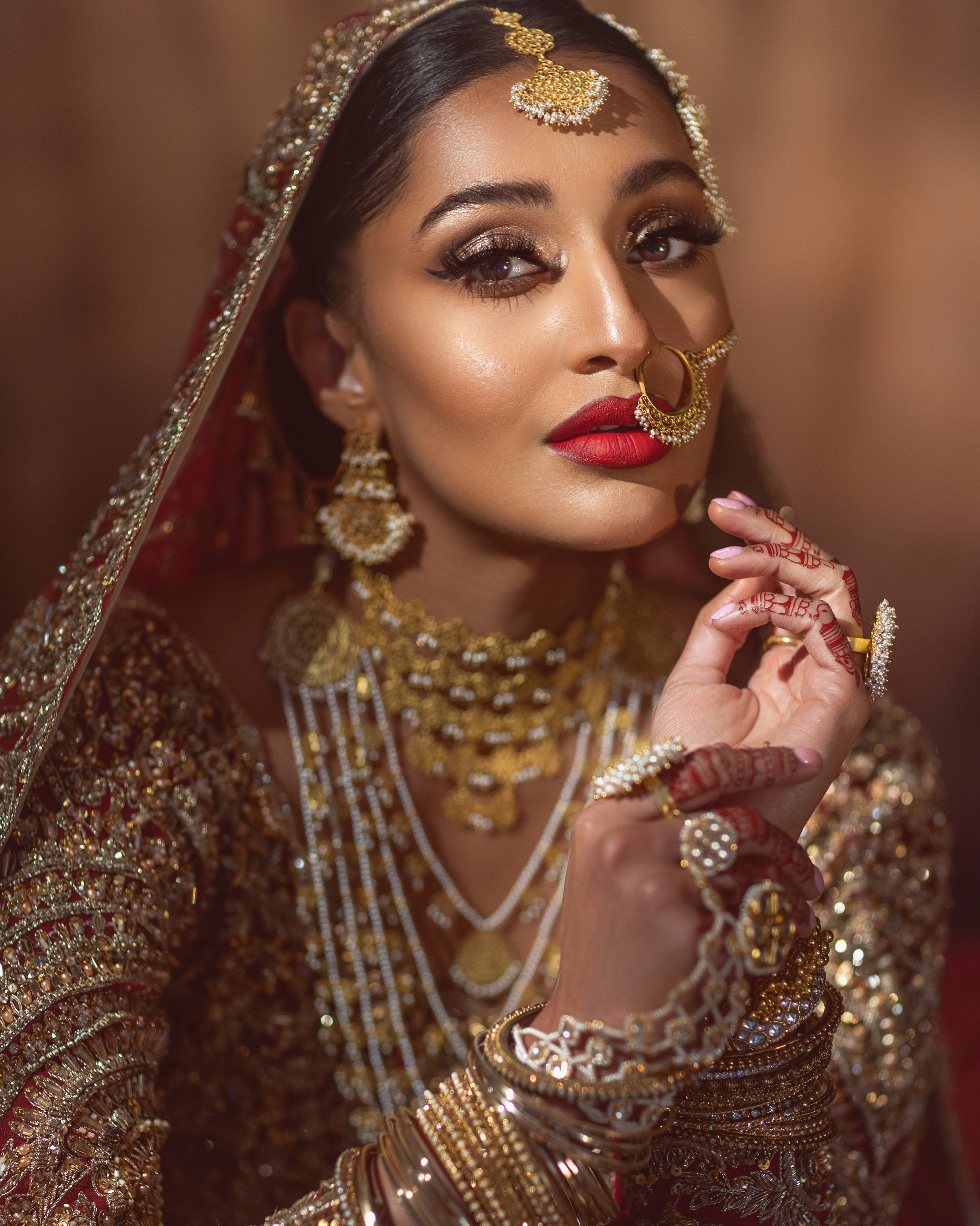 A woman dressed in traditional Indian bridal attire with intricate gold jewelry, including earrings, necklaces, a nose ring, bangles, and rings, with henna designs on her hand and bold red lipstick.