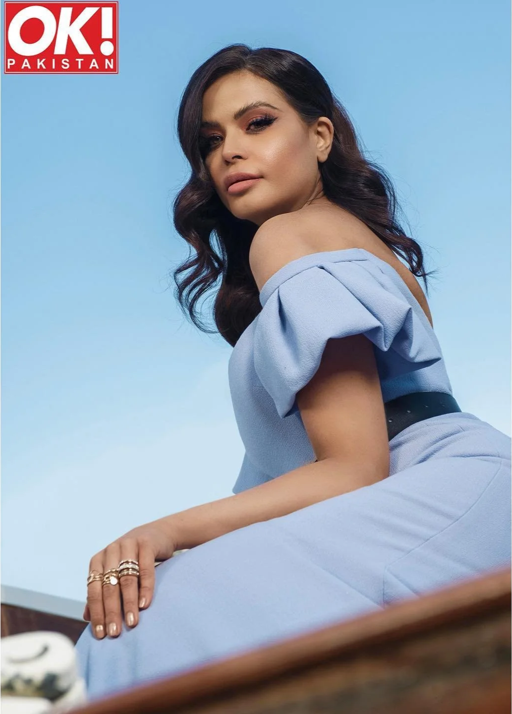 A woman in a light blue off-shoulder outfit sitting outdoors with a clear blue sky background. She has wavy dark hair, makeup, and multiple rings on her fingers. The logo