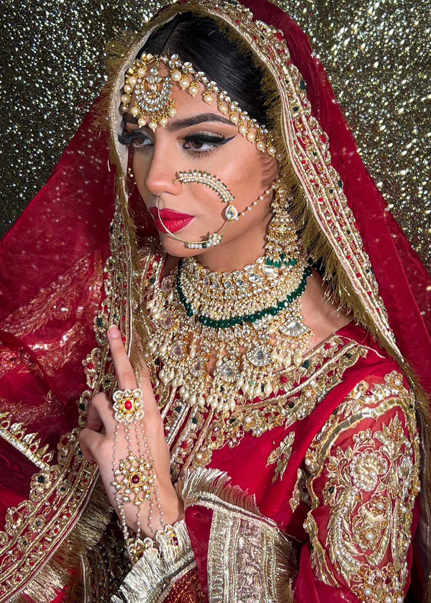 A woman dressed in traditional Indian bridal attire with red and gold embroidered clothing and elaborate jewelry, including necklaces, earrings, and a nose ring, against a glittery background.