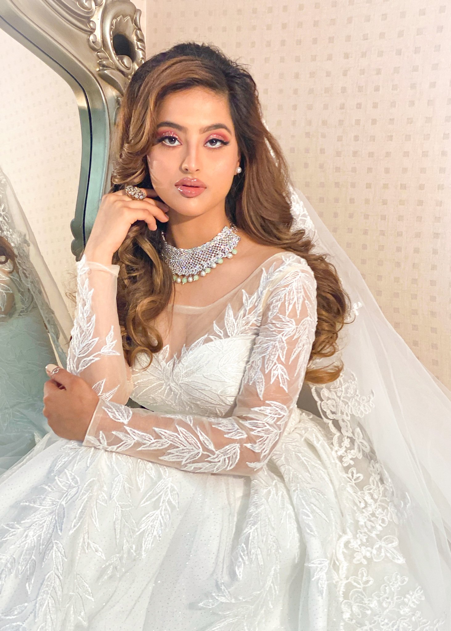 A woman in a wedding dress with floral embroidery, wearing jewelry including a pearl necklace, earrings, and rings, sitting on a white chair near a mirror with ornate detailing.