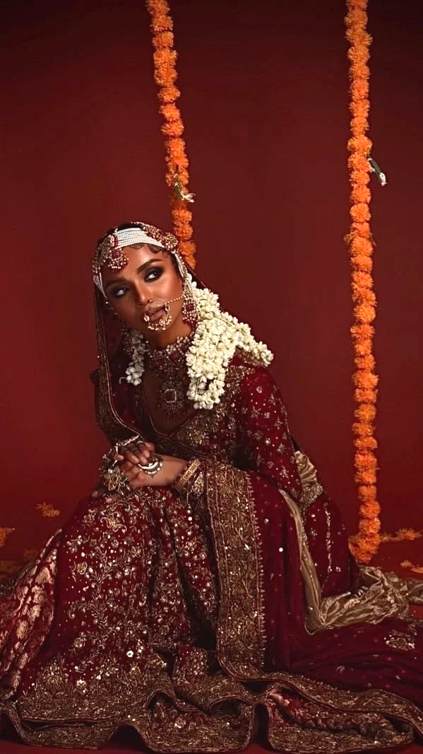 A woman dressed in traditional Indian bridal attire, sitting against a maroon background decorated with orange marigold flower garlands.