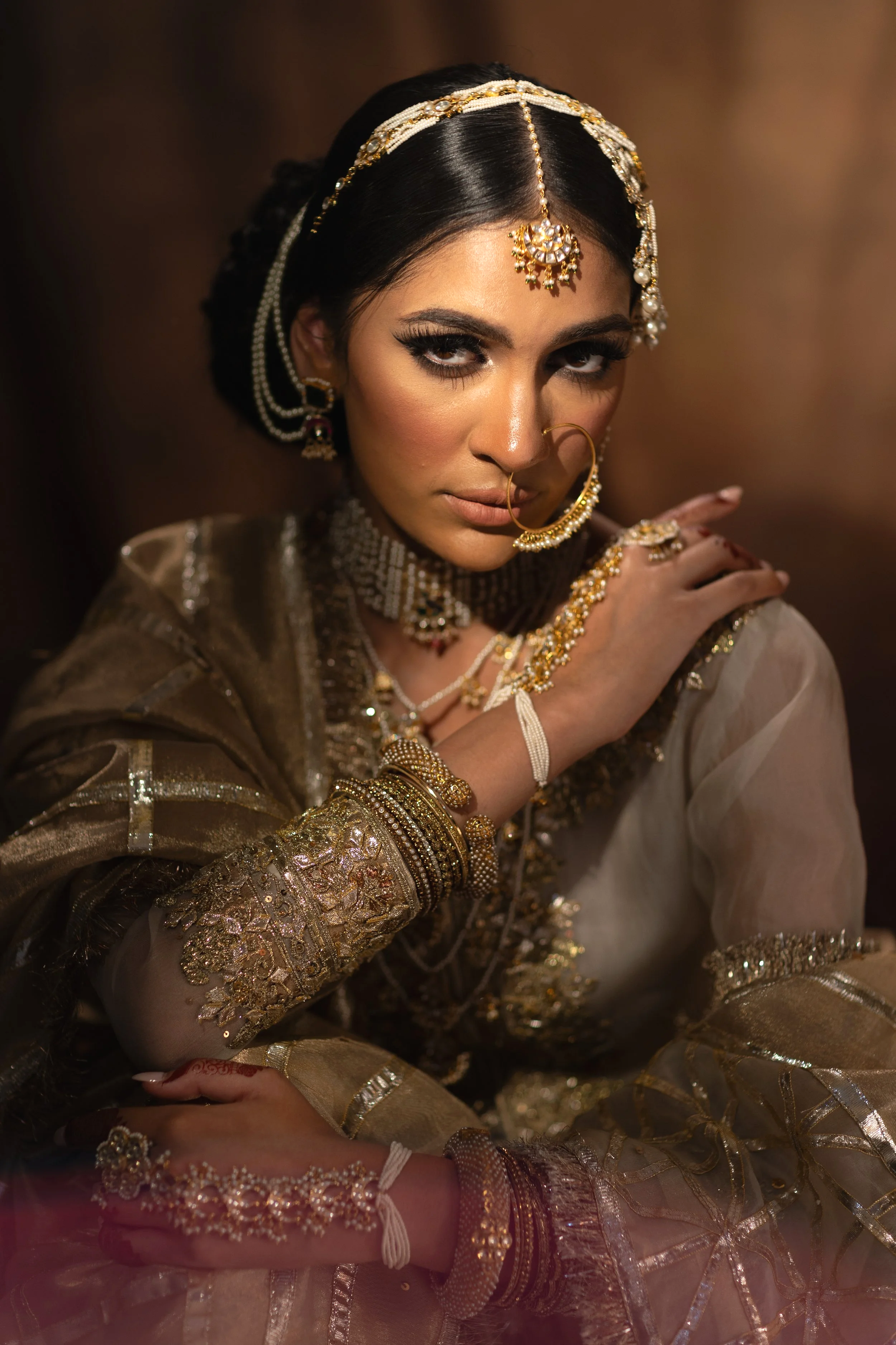 A woman dressed in traditional Indian bridal attire, adorned with gold jewelry including a headpiece, earrings, nose ring, necklaces, and bangles, posing with a confident expression.