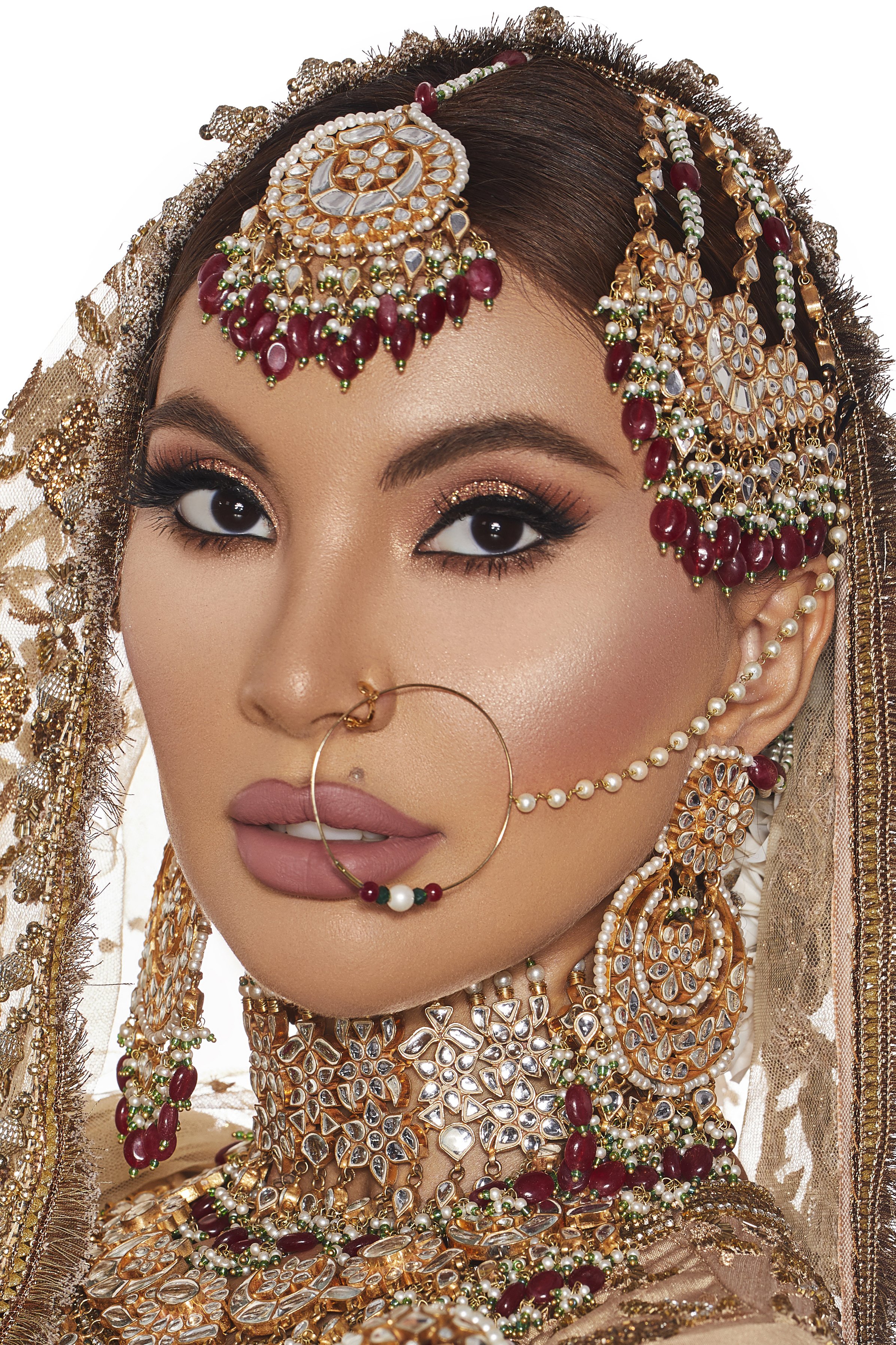 A woman wearing traditional Indian jewelry, including a large nose ring connected to her ear with a chain, intricate earrings, and a heavily decorated head covering with jewelry, pearls, and gemstones.