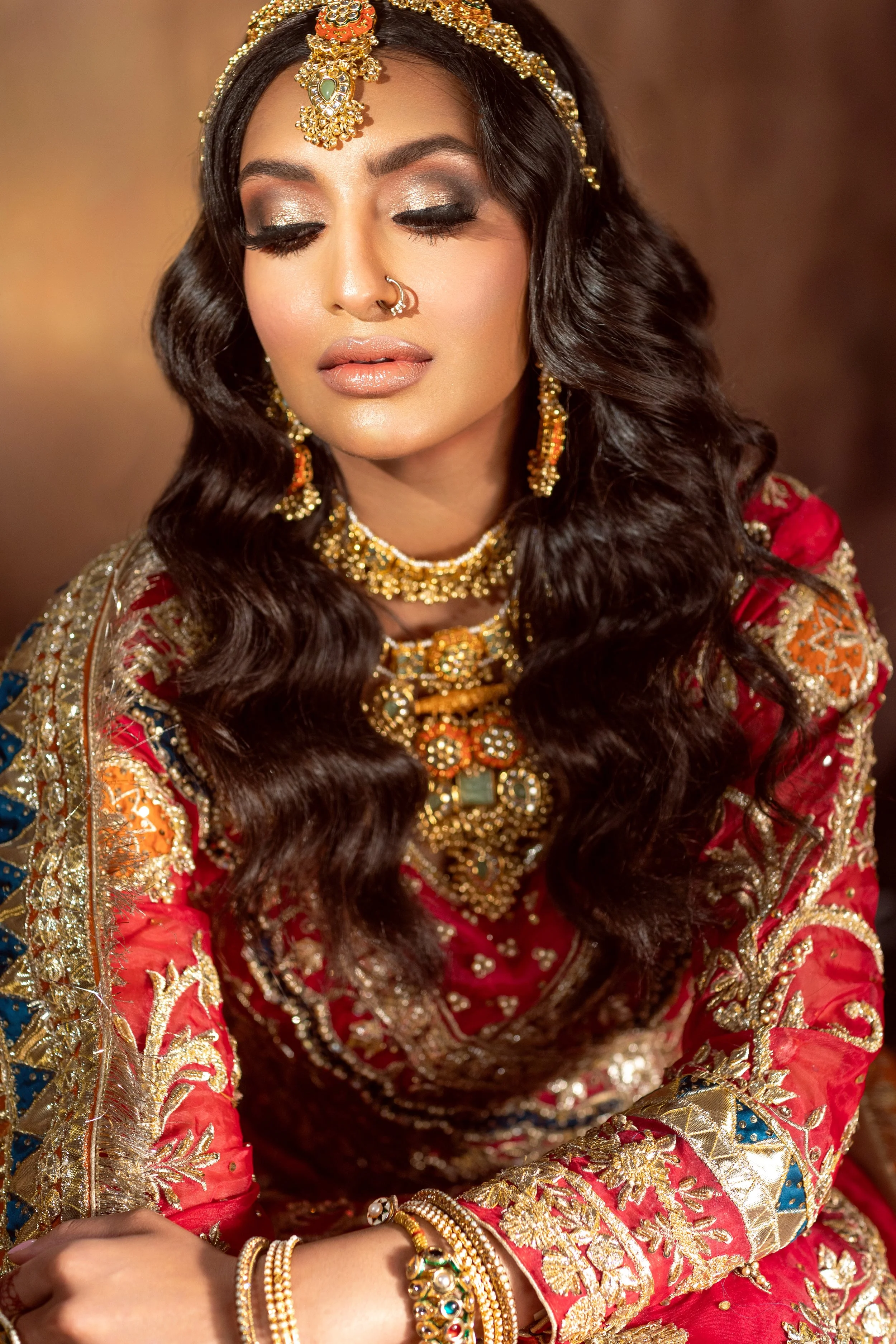 A woman with long, wavy dark hair in traditional Indian attire, wearing elaborate gold and gemstone jewelry, including a headpiece, earrings, necklace, and bangles, with her eyes closed, in front of a warm background.
