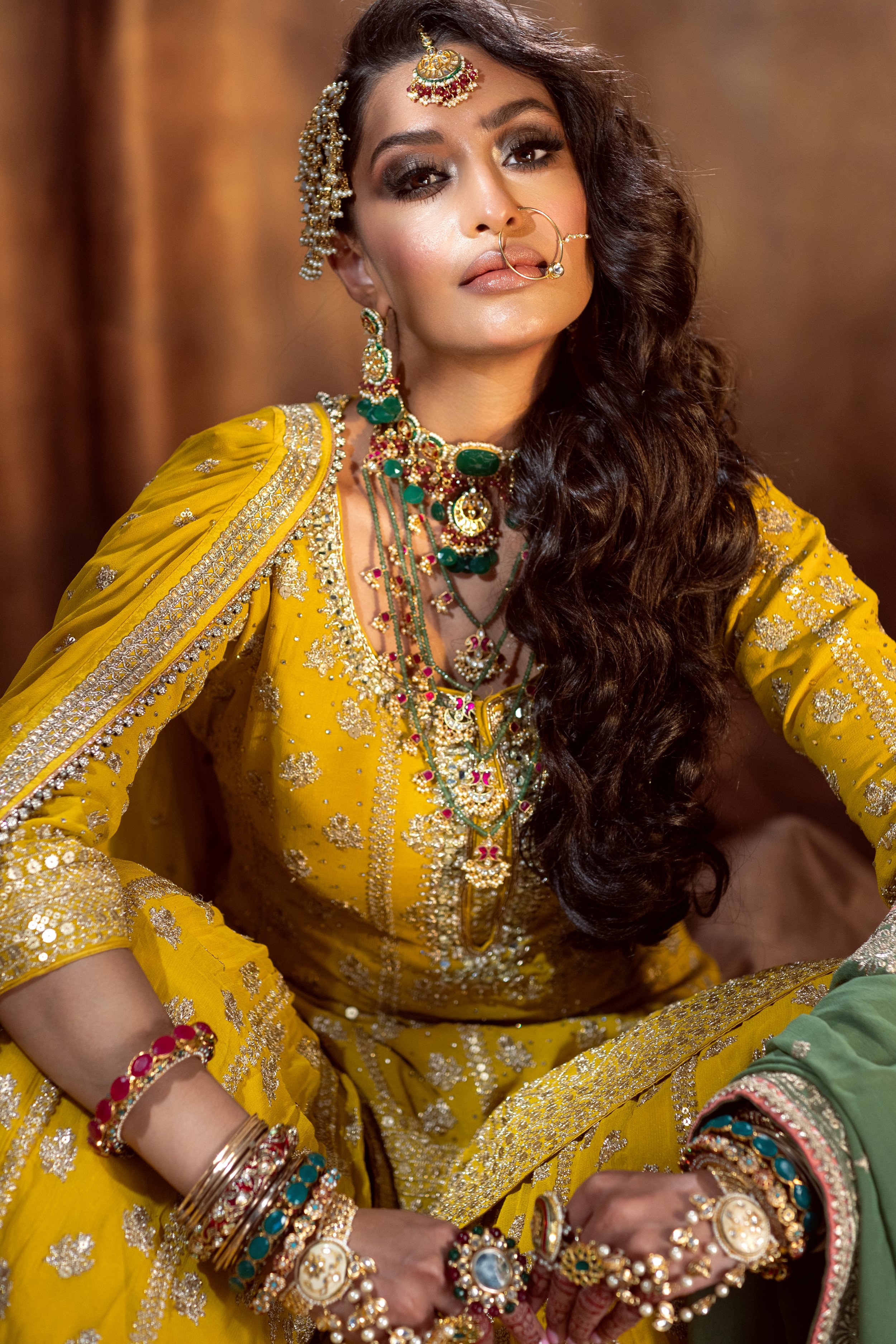 A woman dressed in a traditional yellow Indian outfit adorned with gold embroidery and jewelry, including necklaces, earrings, bangles, a nose ring, and a maang tikka, sitting with her arms resting on her lap, against a warm brown background.