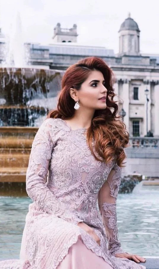 Woman with long, wavy auburn hair in a light pink lace dress, sitting near a fountain in a cityscape background with historic buildings.