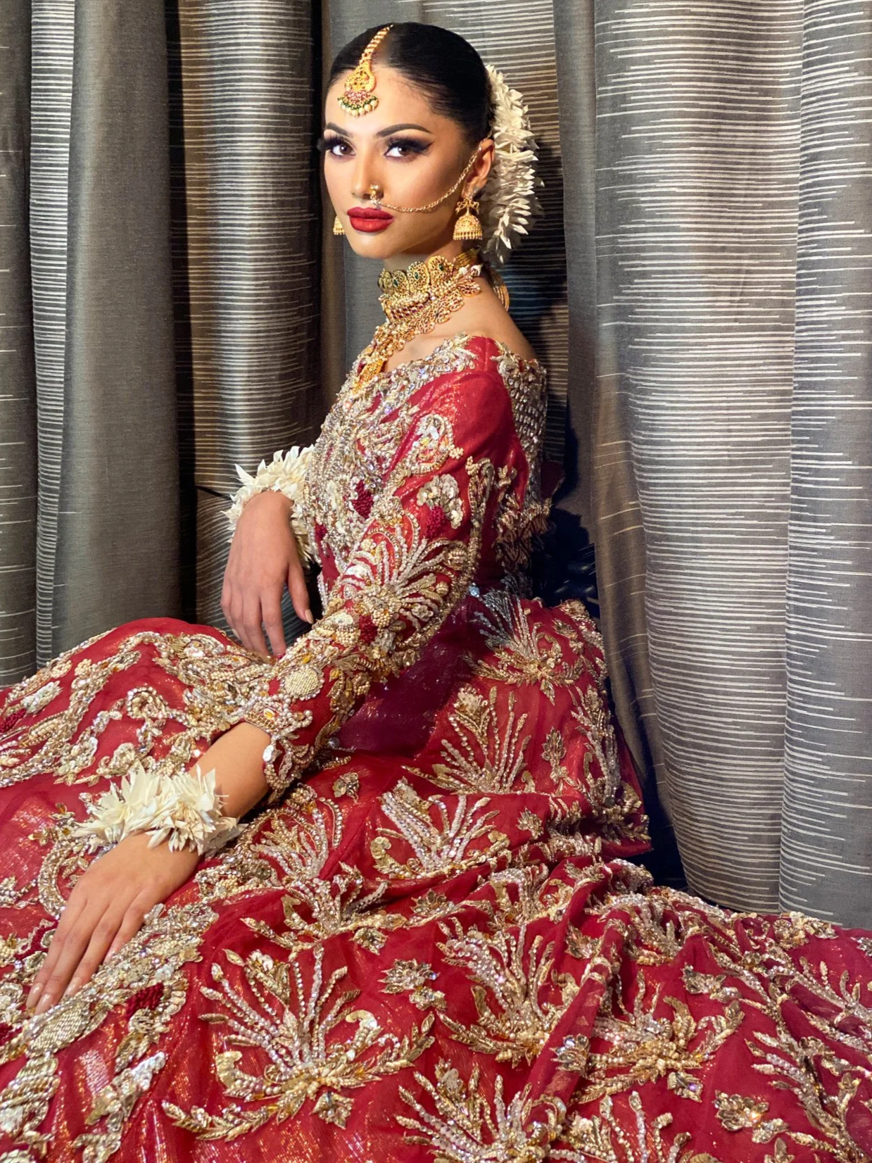 A woman in traditional Indian attire with intricate gold embroidery, jewelry, and flowers in her hair, sitting against a curtain background.