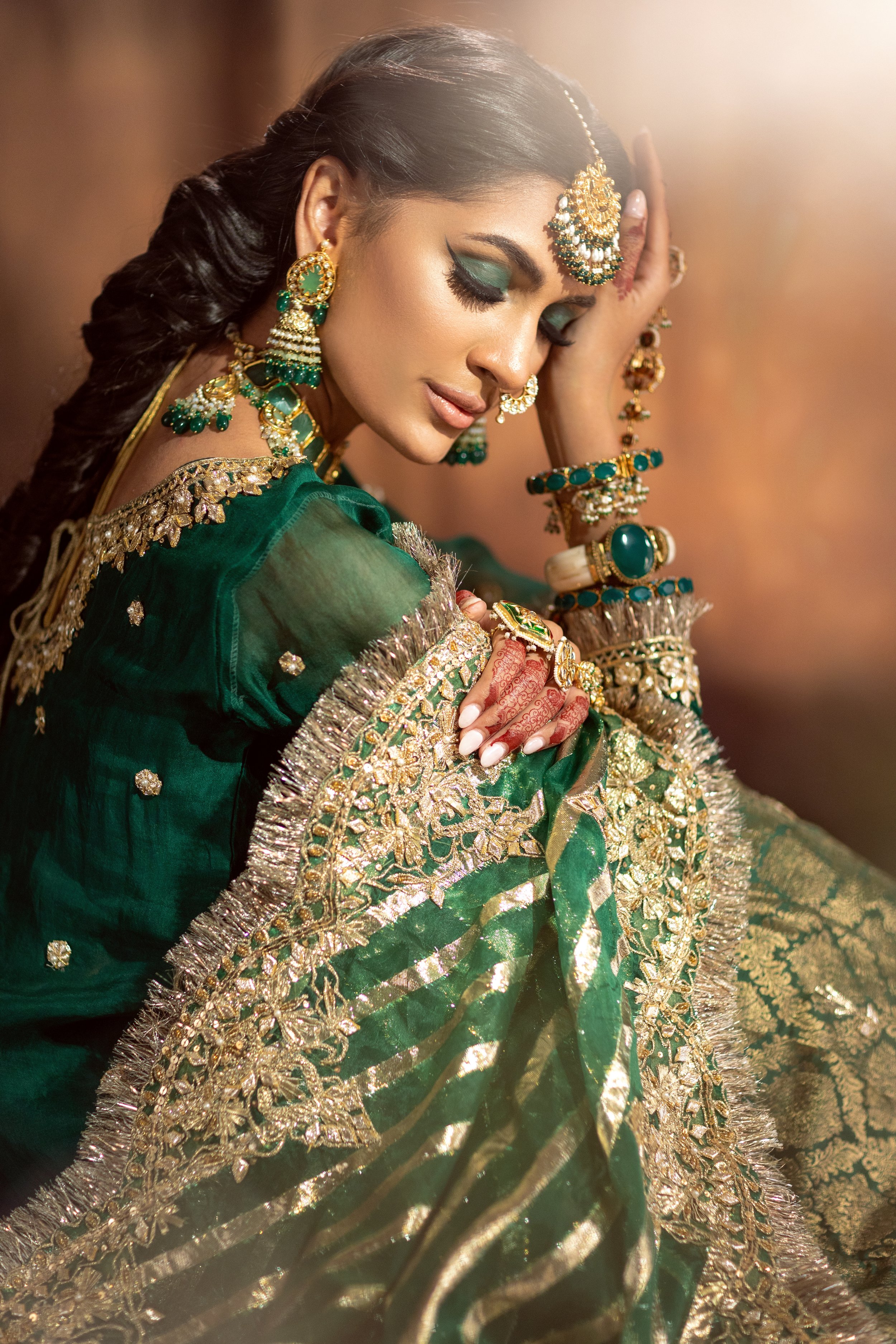 A woman dressed in traditional Indian attire, wearing elaborate gold and green jewelry, including earrings, necklaces, and bangles, with henna on her hands and vibrant makeup, sitting with her eyes closed and hand on her forehead.