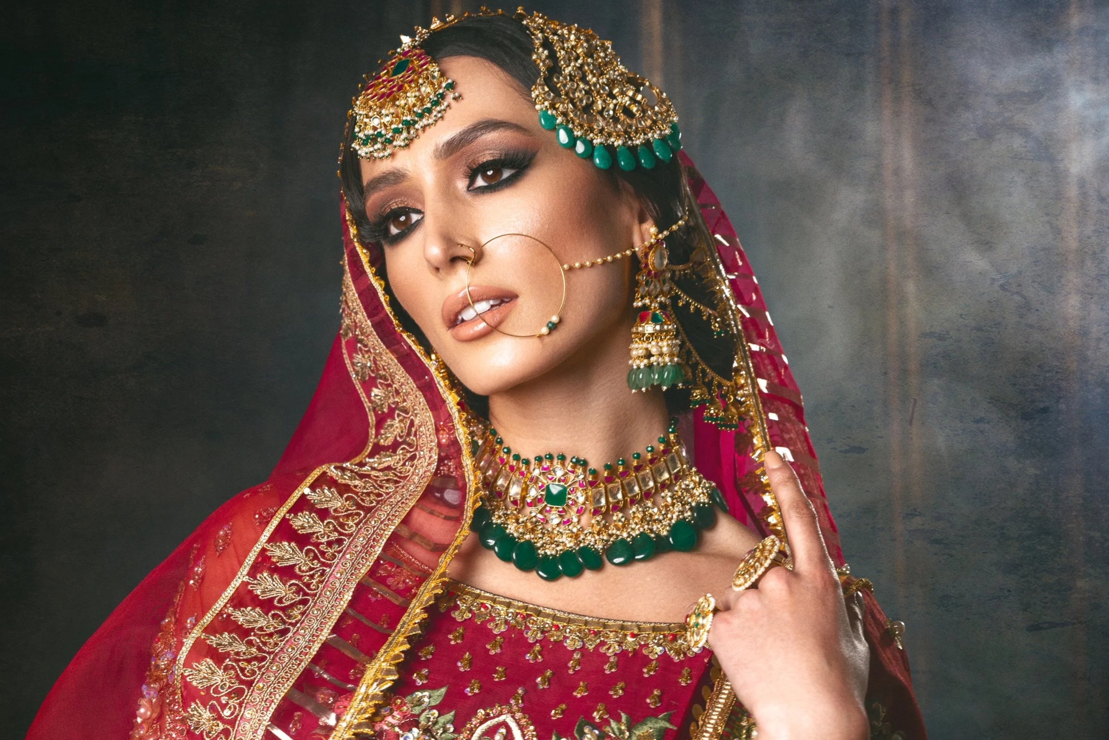 A woman dressed in traditional Indian bridal attire with intricate gold jewelry, including a maang tikka, nose ring, earrings, necklace, and rings. She wears a red and gold embroidered saree with a matching red veil.