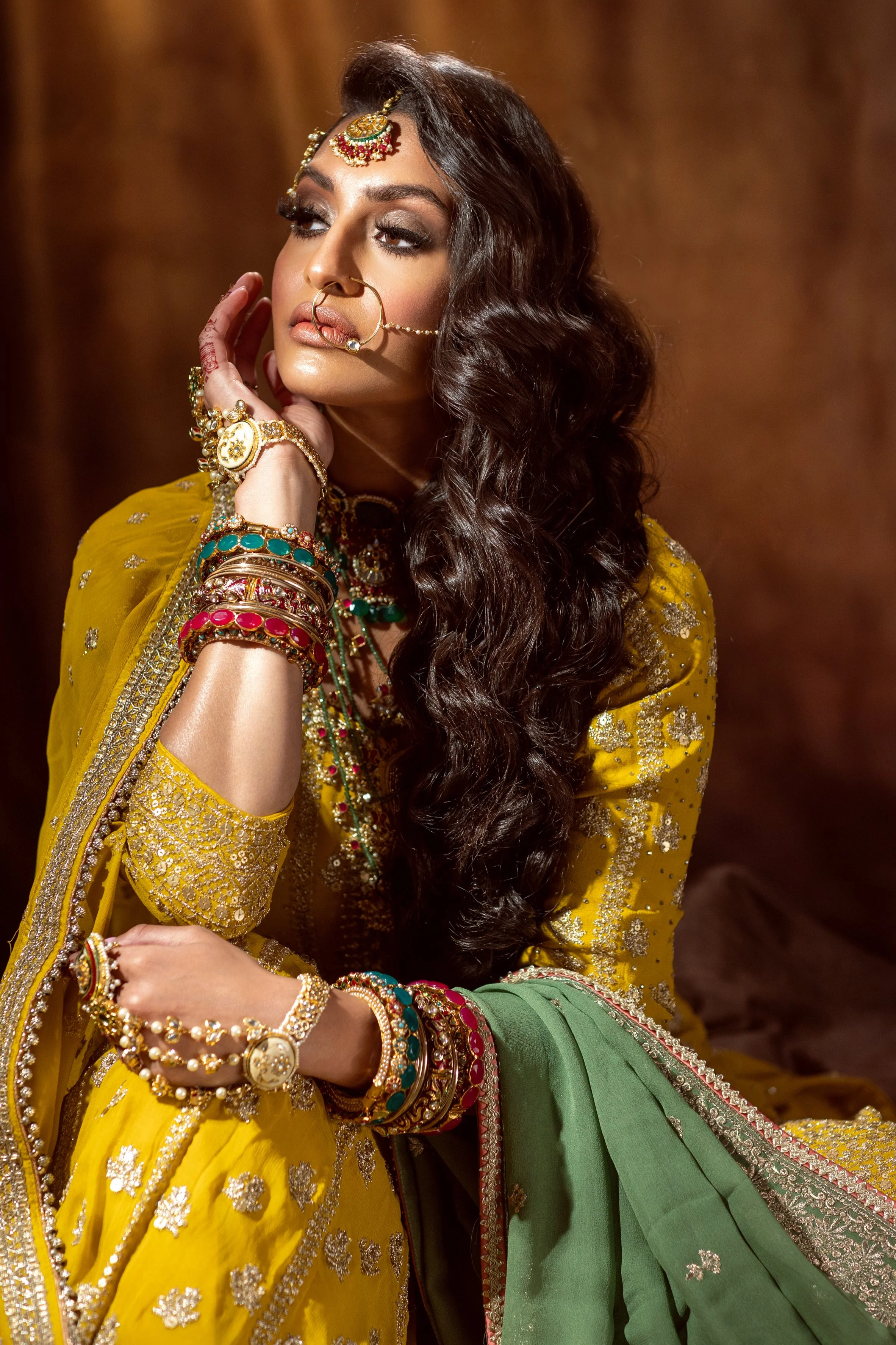 A woman dressed in traditional Indian attire with elaborate jewelry, including necklaces, bangles, earrings, and a nose ring, posed against a warm-colored background.