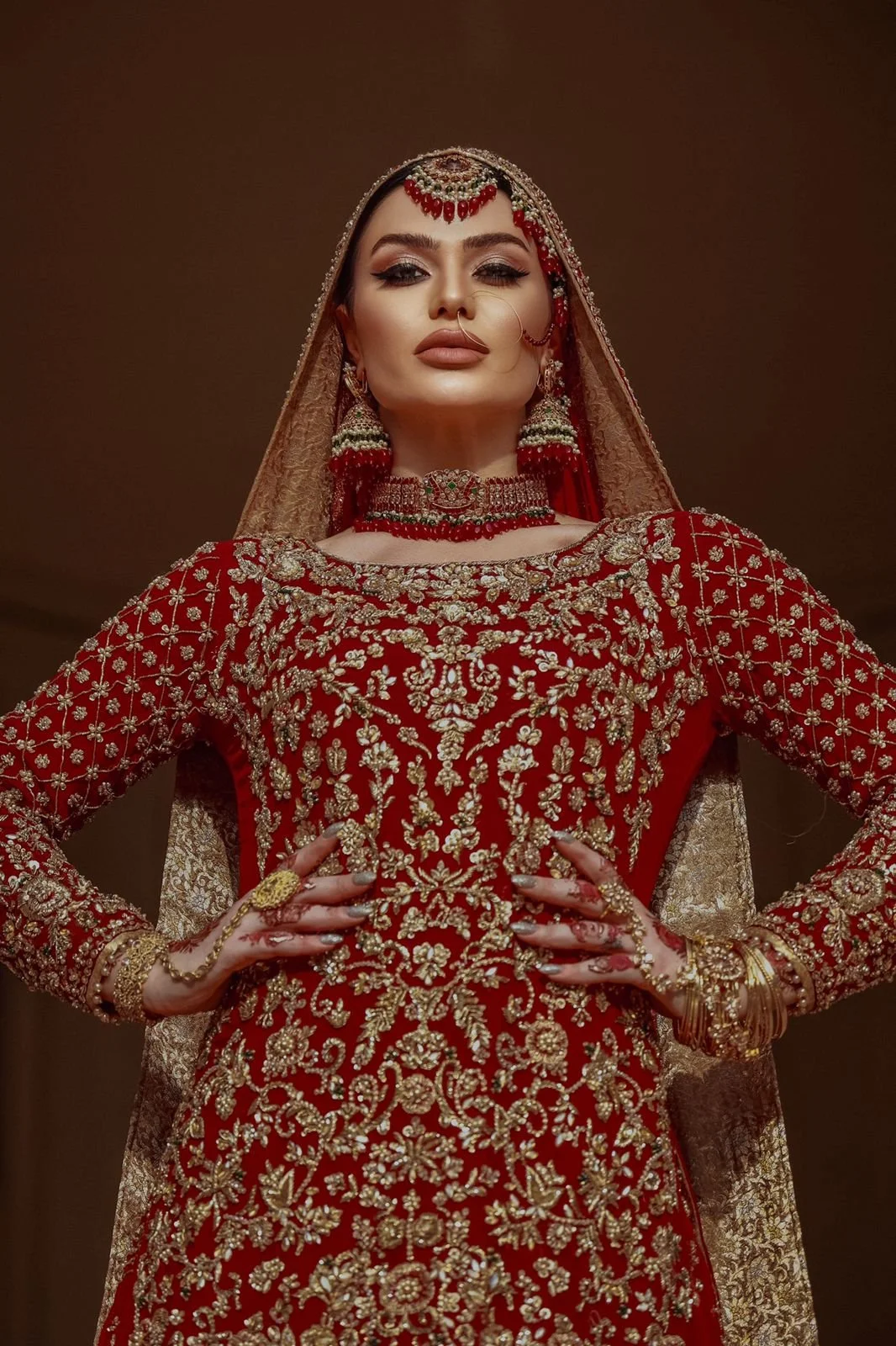 A woman dressed in traditional Indian bridal attire, wearing a red and gold embroidered gown, jewelry, and a veil.