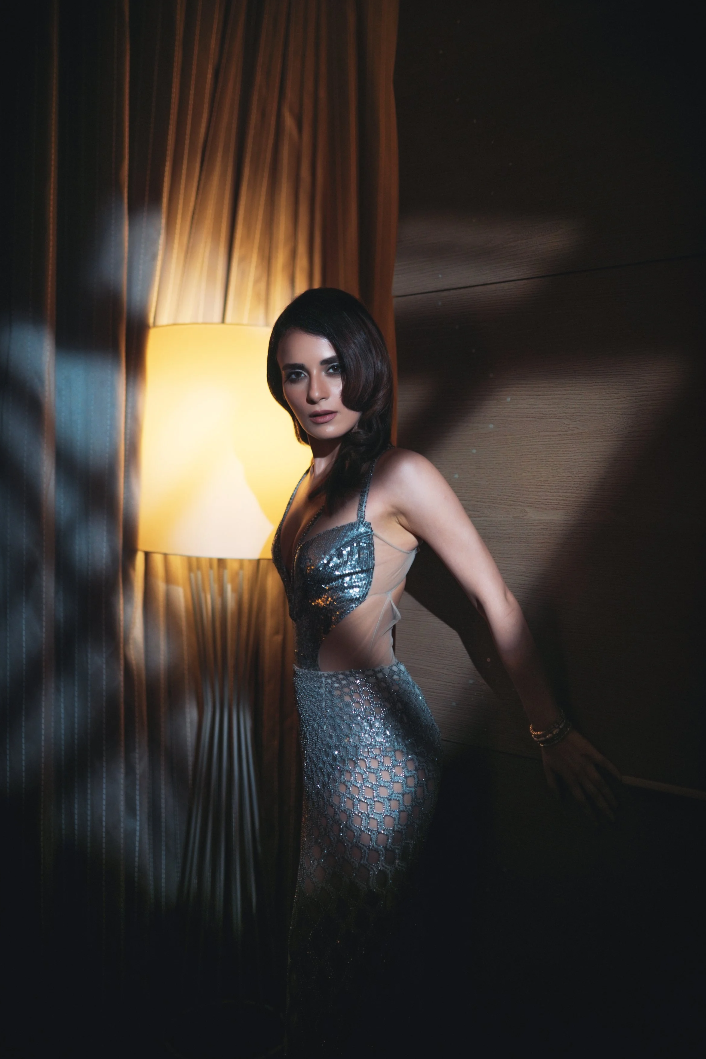 A woman with dark hair and fair skin in a glamorous silver, eye-catching dress, standing by a wooden wall next to a lit lamp in a dimly lit room.