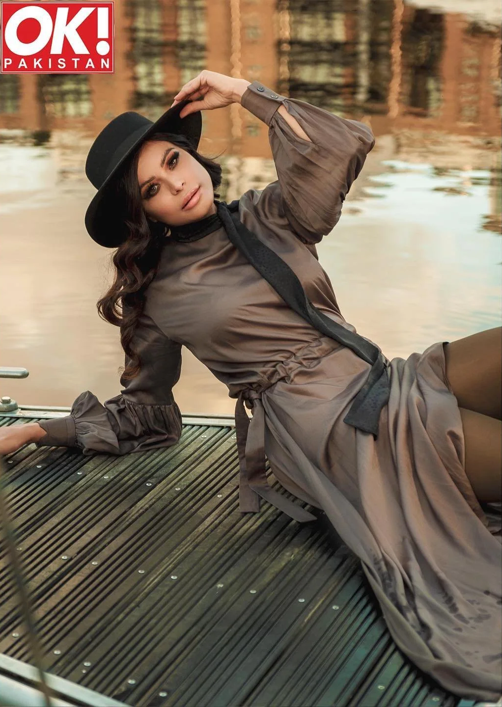A woman in a brown dress and black hat sitting on a dock by the water, with reflections of buildings in the background.