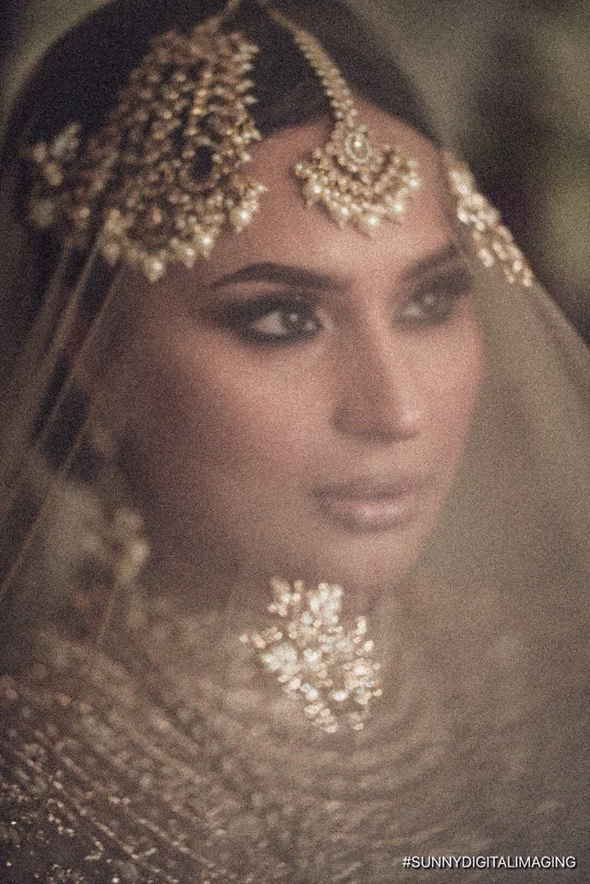 Close-up of a woman wearing traditional Indian bridal jewelry, including a forehead ornament and necklace, with detailed makeup and a veil.