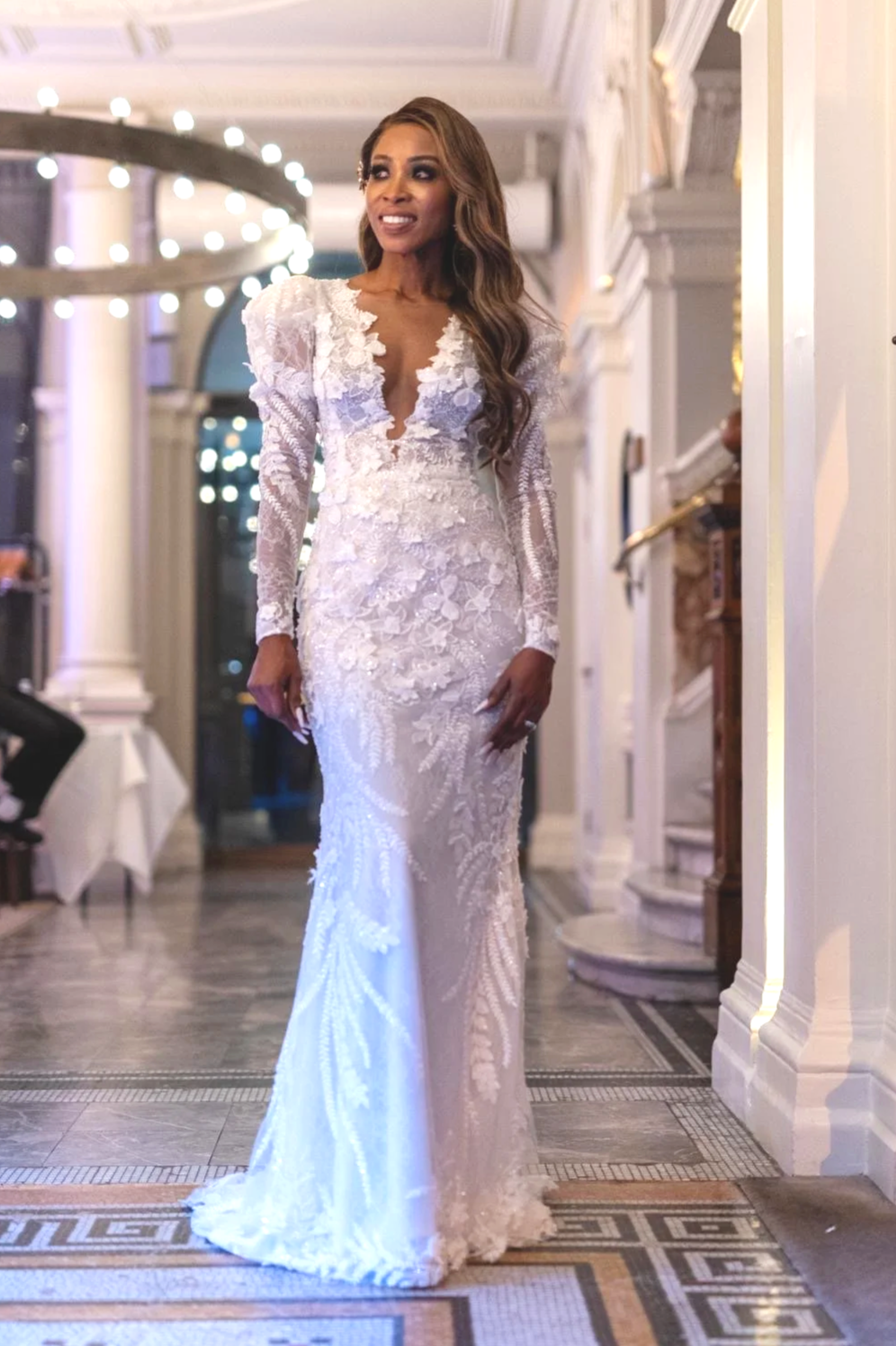 A woman in a white, lace wedding dress with long sleeves and floral embroidery, standing in an elegant interior with chandeliers and ornate decor.