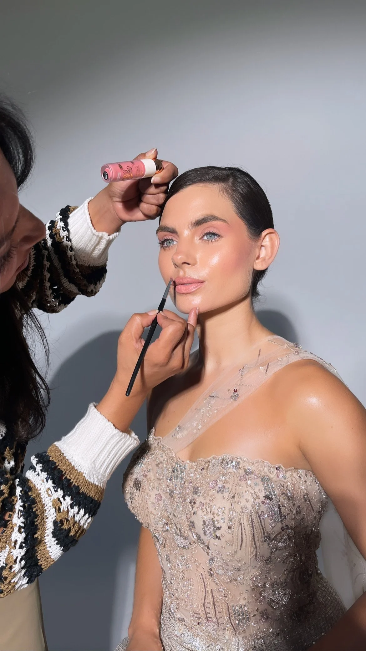 A woman with short black hair and wearing an elegant, embellished dress is having her makeup touch-up done by a makeup artist. The makeup artist is applying lipstick with a brush and holding a pink makeup product.