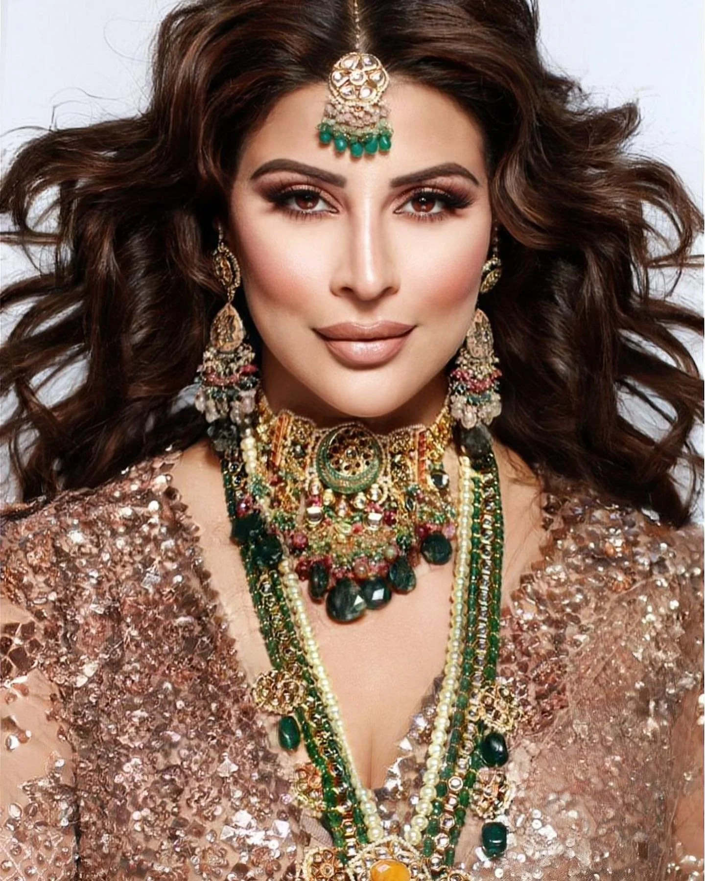A woman with long curly hair wearing traditional jewelry, including earrings, necklaces, and a headpiece, dressed in a sequined gown.
