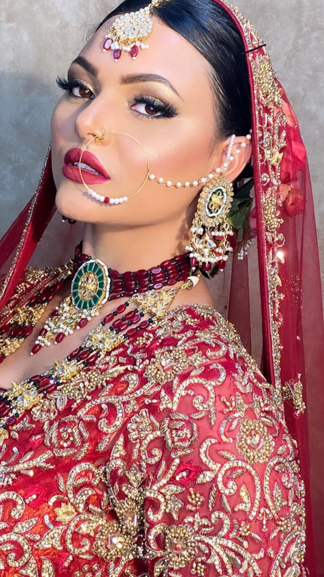 A woman wearing traditional Indian wedding attire with red and gold embroidery, adorned with elaborate jewelry including earrings, necklace, nose ring, and forehead ornament, and a sheer red veil with gold embellishments.