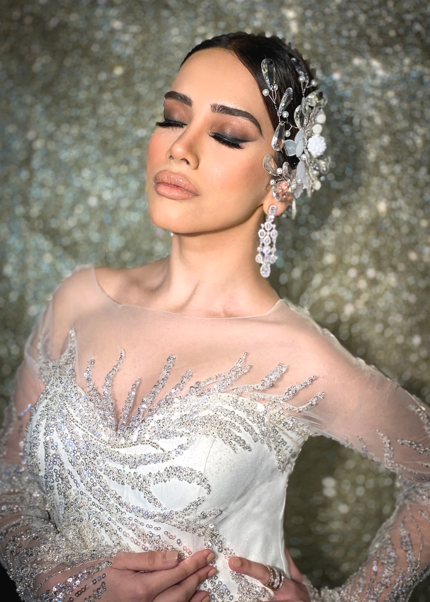 A woman with her eyes closed, wearing makeup and large earrings, with her hair decorated with a floral and gem headpiece, dressed in a white dress with beadwork, standing in front of a sparkly gold background.