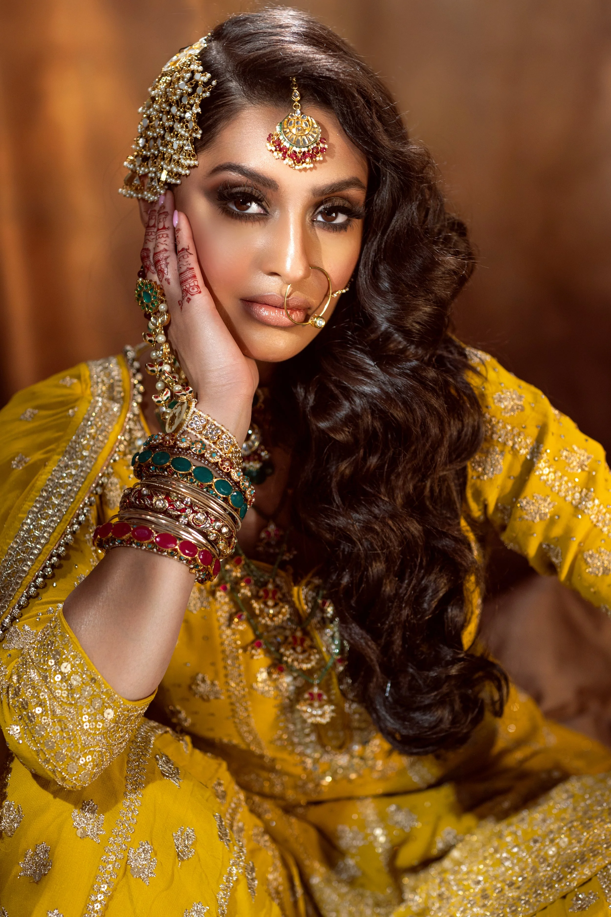 A woman dressed in traditional Indian attire, wearing elaborate jewelry and henna on her hand, posing with her hand near her face.