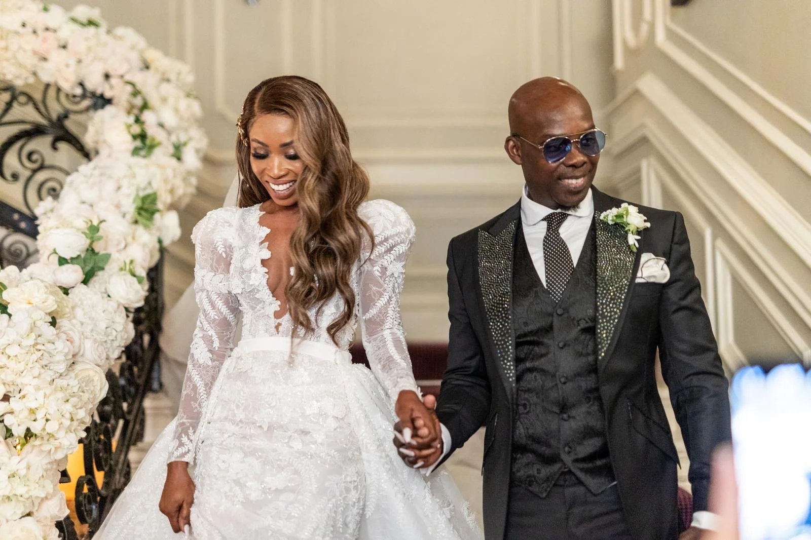 A newly married African American couple, the bride in a white wedding gown and the groom in a black suit with sunglasses, holding hands and smiling inside a decorated venue.