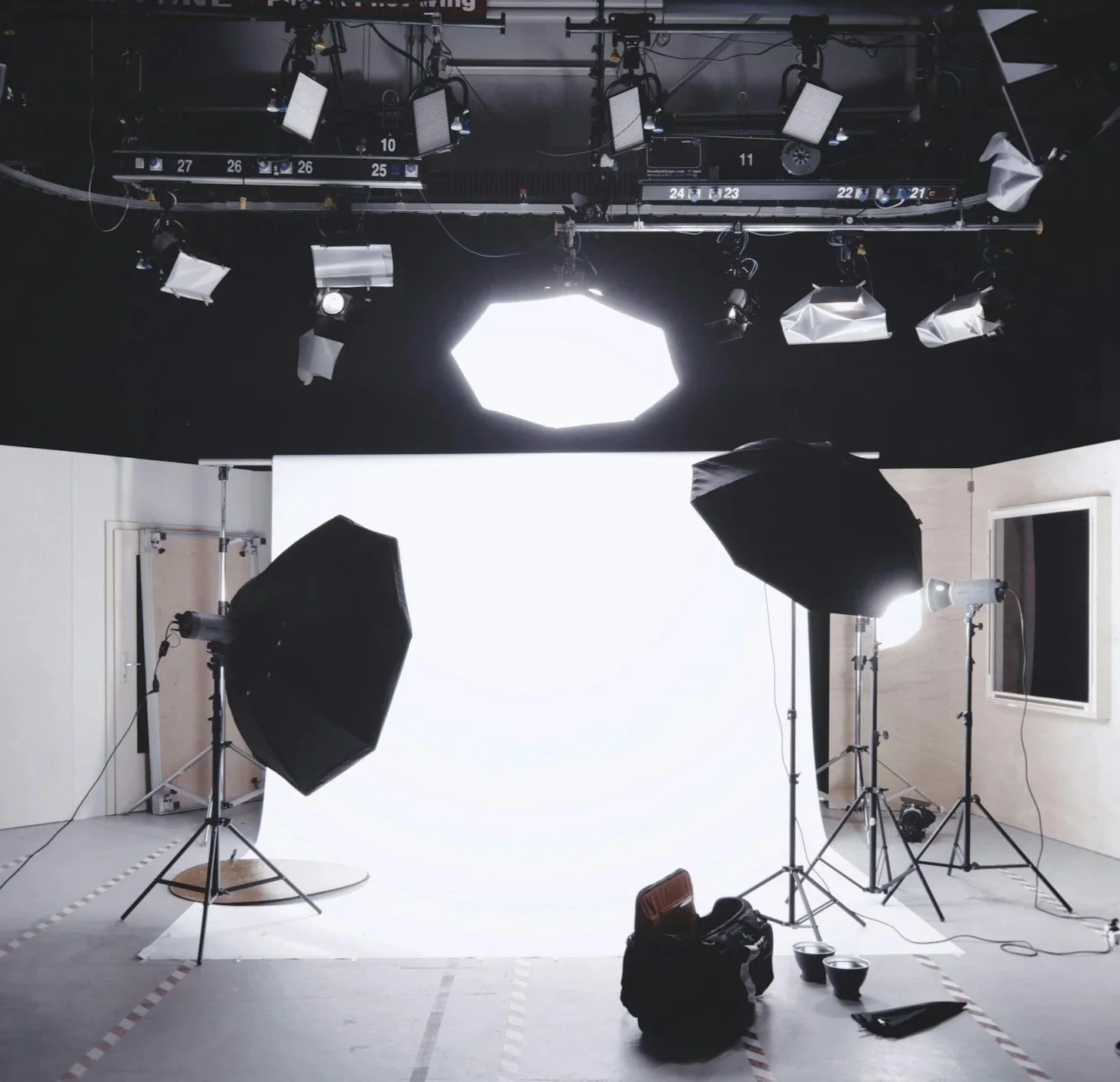 A professional photography or video studio set up with multiple studio lights and umbrellas directed at a white backdrop, camera equipment, and a door on the wall.