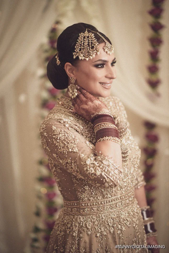 A woman dressed in traditional Indian bridal attire, wearing ornate jewelry and henna on her hands, smiling with her eyes closed, in a decorated setting.